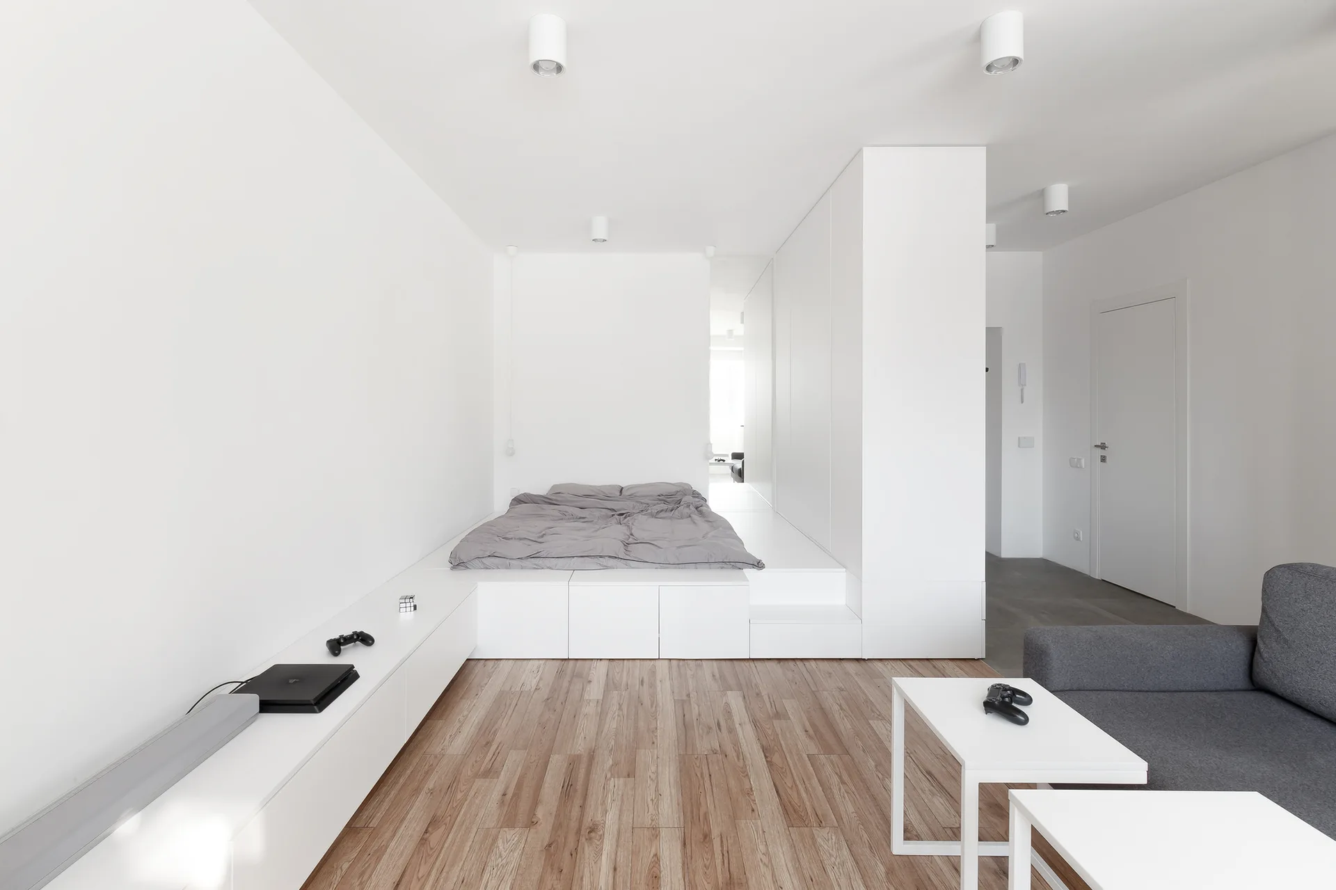 A minimalist studio apartment features a built-in white platform bed, a low storage console, light wood floors, and a grey sofa.