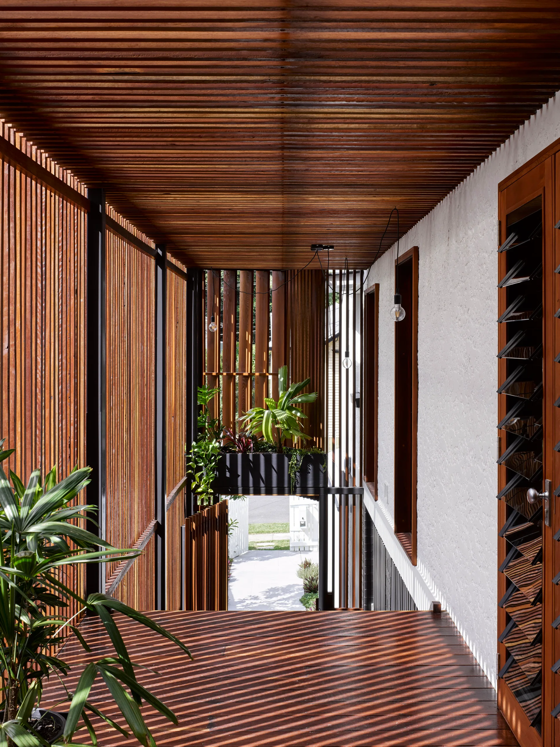 A semi-enclosed outdoor walkway features vertical wooden slats on the walls and ceiling casting striped shadows across a dark wood deck.