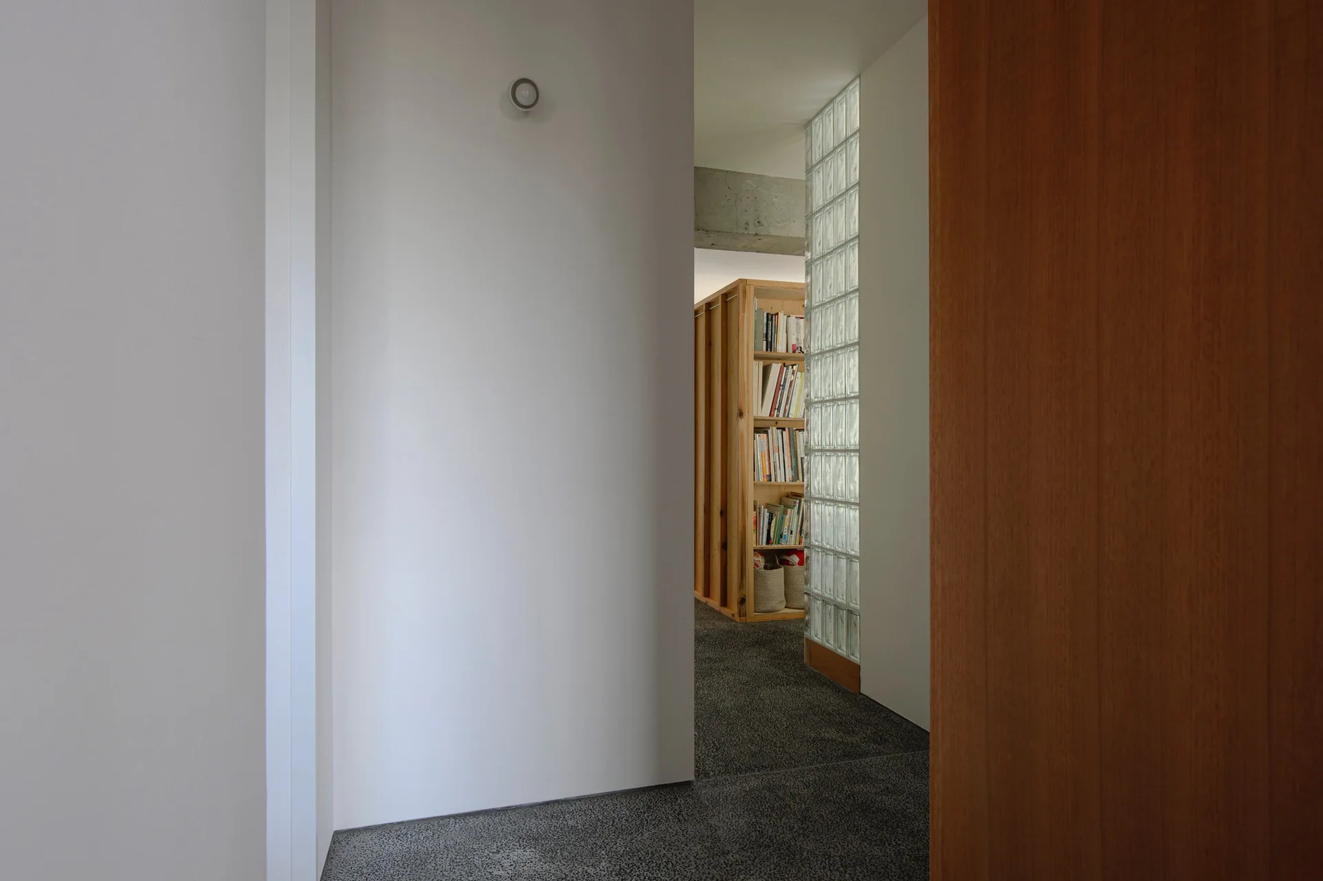 A view through a doorway into a minimalist hallway featuring a vertical glass block partition, a wooden bookcase, and an exposed concrete ceiling beam.