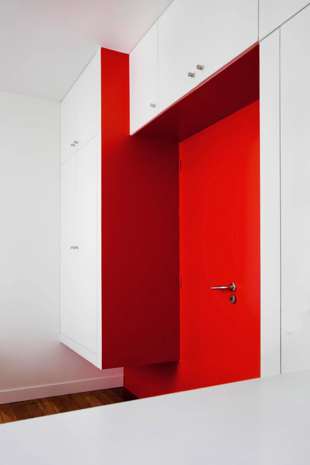 A modern hallway features a recessed vibrant red doorway set within a wall of minimalist matte white built-in storage cabinets.