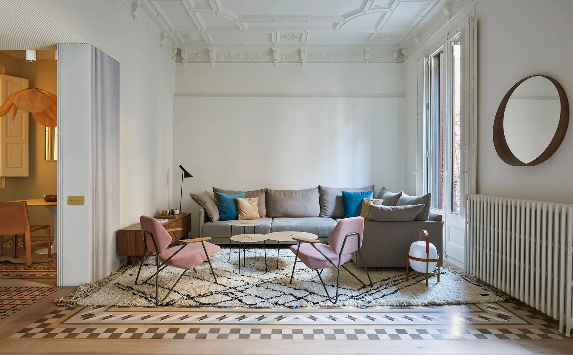 A living room with an ornate plaster ceiling features a gray sectional sofa, two pink armchairs, and a patterned rug on a decorative tile floor.