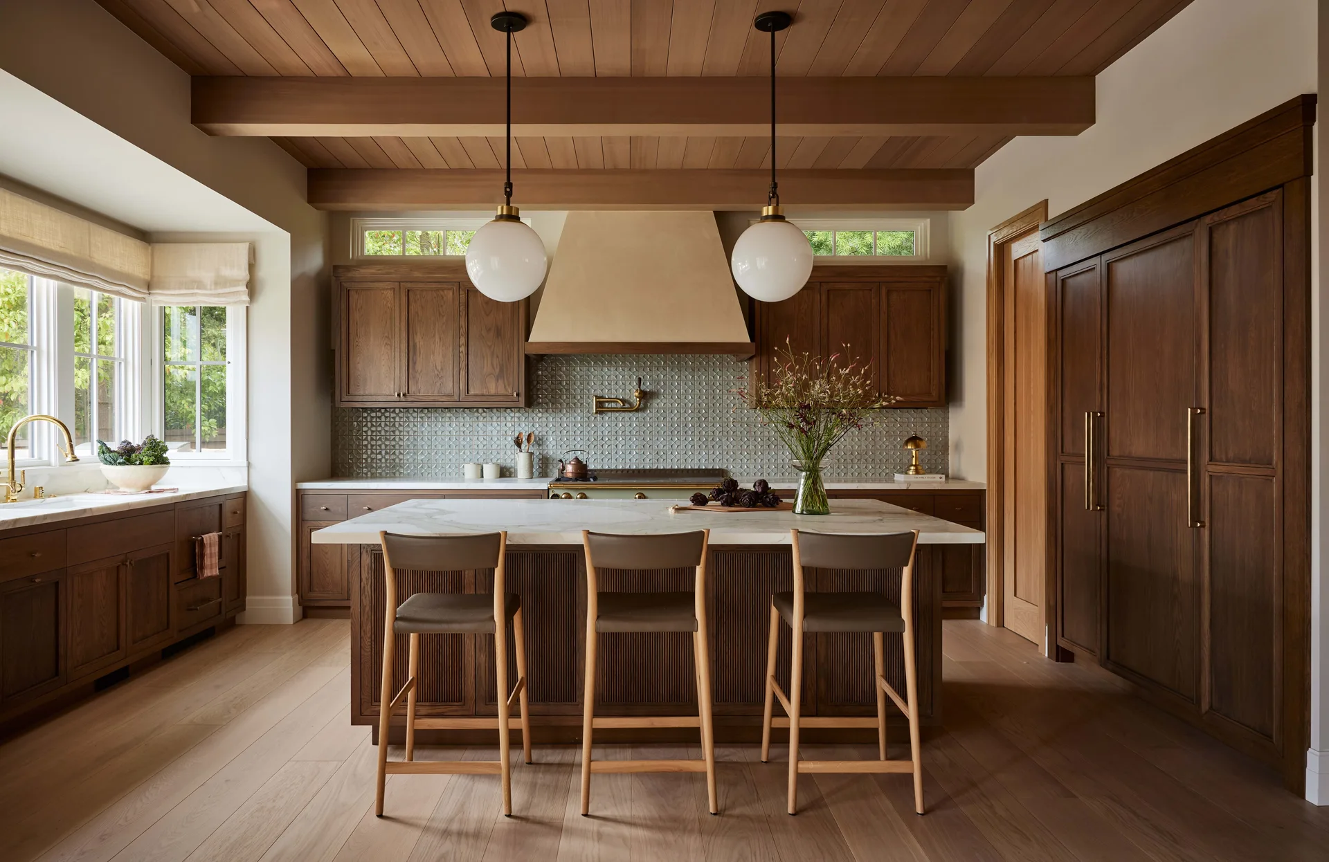 A spacious kitchen features warm wood cabinets, a plaster range hood, a marble-topped island with three bar stools, and globe pendant lights.