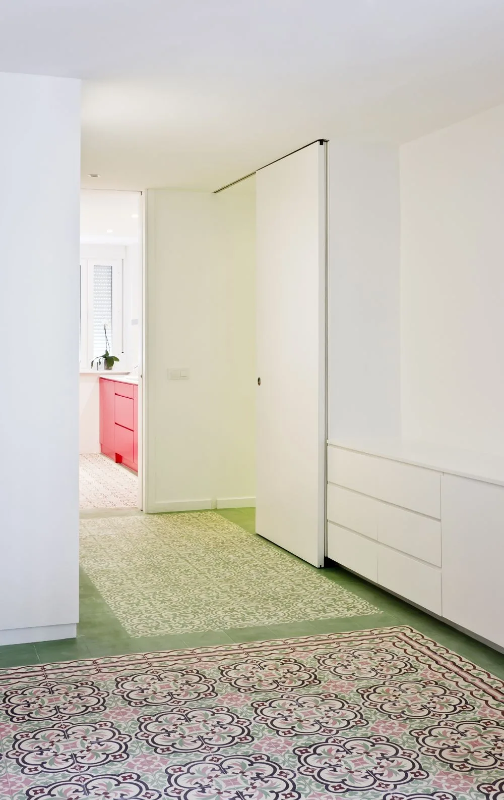 A minimalist white hallway featuring patterned green and pink floor tiles, white built-in drawers, and a view of pink kitchen cabinets.