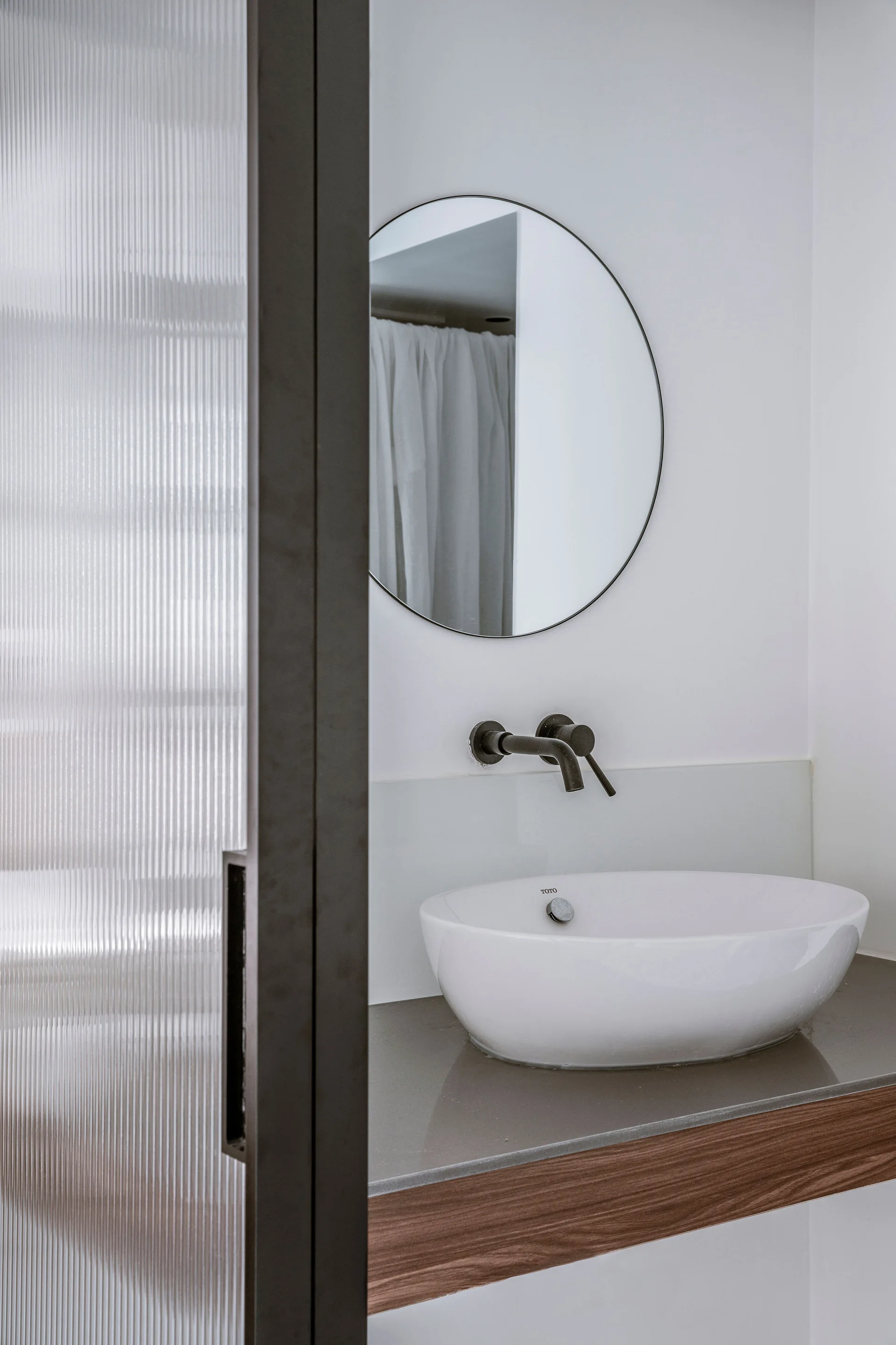 A white vessel sink rests on a grey countertop beneath a round mirror and matte black faucet, partially framed by a fluted glass partition.