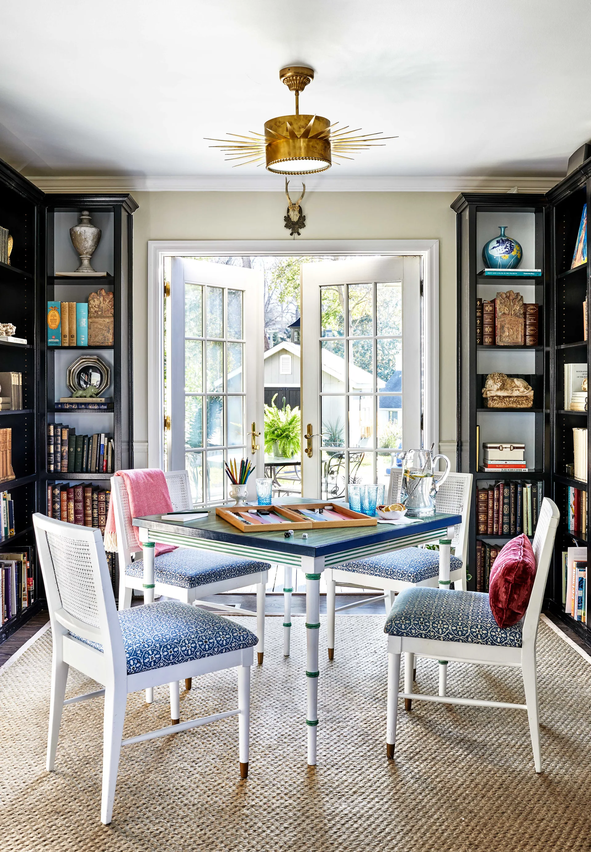 A home library featuring a central game table with four blue patterned chairs, flanked by tall black bookshelves and open French doors.