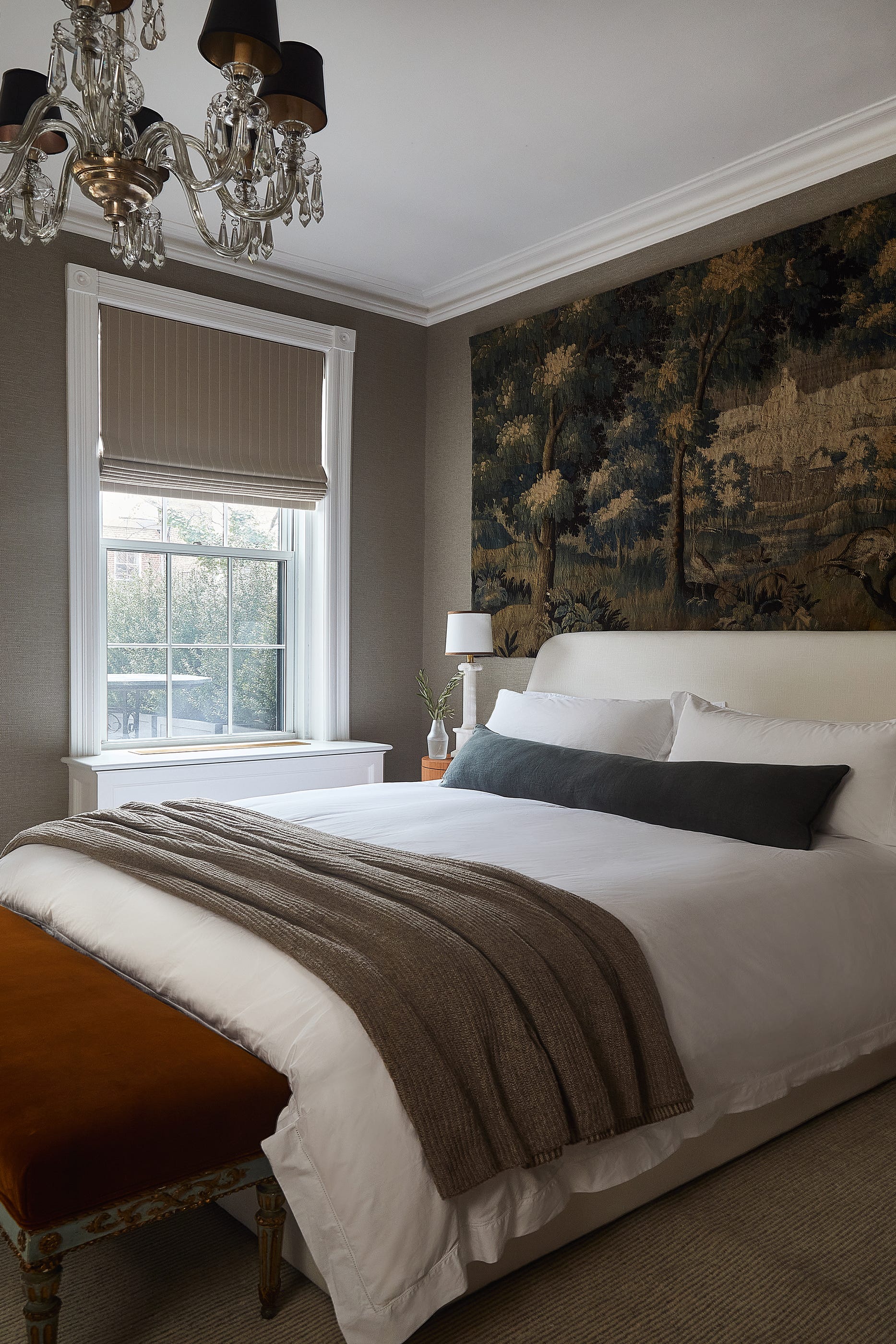 A bedroom features an upholstered bed with a large woodland tapestry on the wall, a window with a roman shade, and a crystal chandelier.