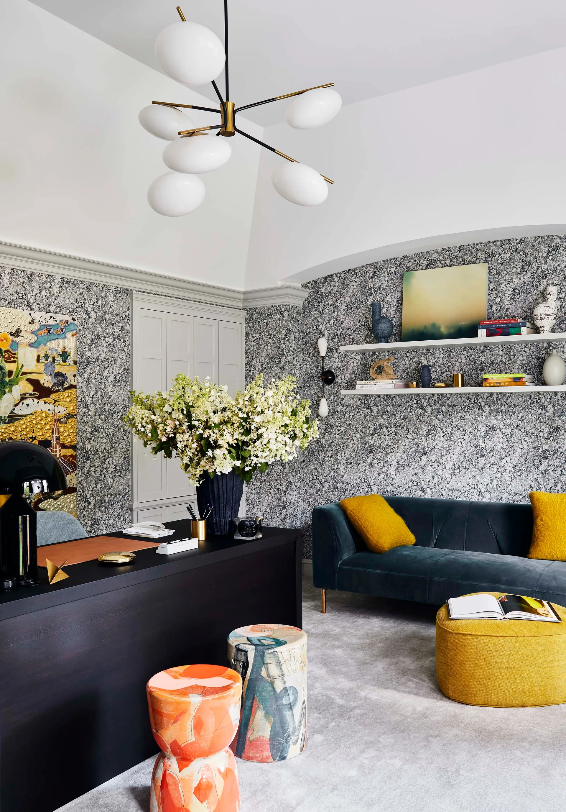 A home office features grey marbled wallpaper, a black desk with patterned stools, and a blue velvet sofa with yellow accent pillows.