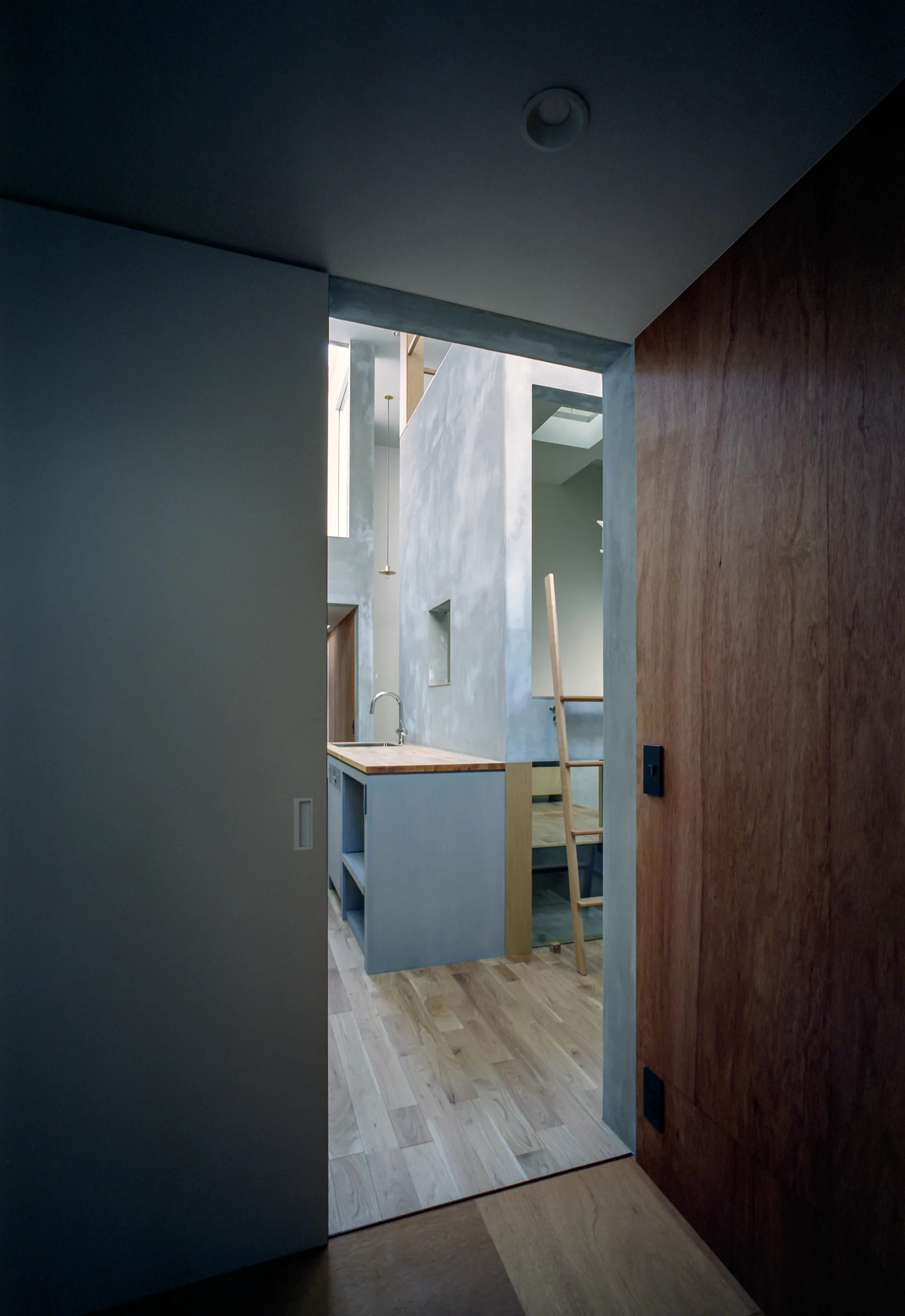A view through a sliding doorway showing a minimalist grey kitchen island with a wood countertop and grey plaster walls.