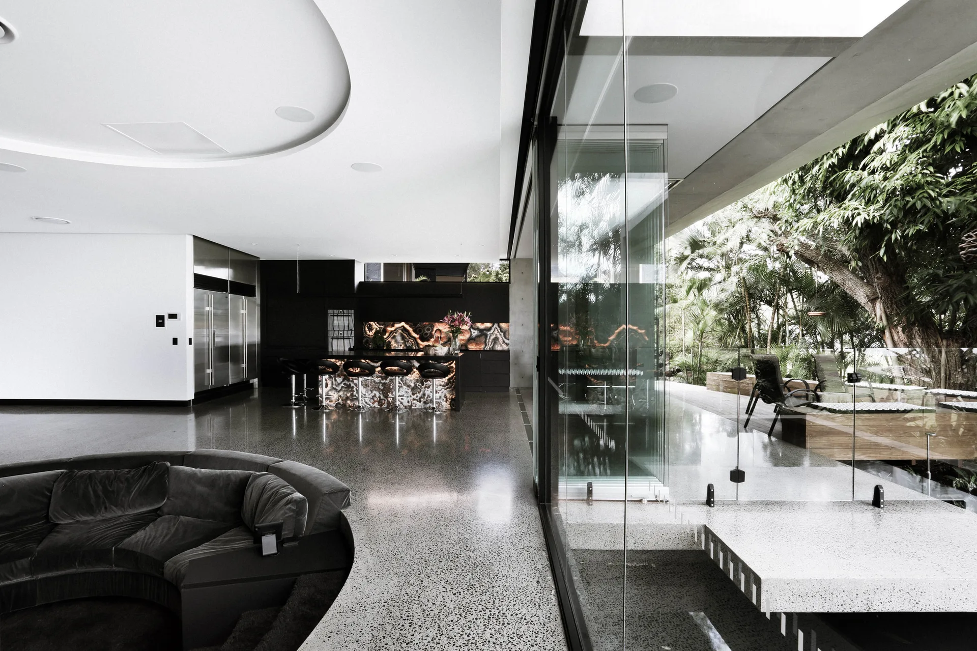 A spacious modern interior showing a backlit stone kitchen island, a curved dark sofa, and large sliding glass doors leading to a patio.