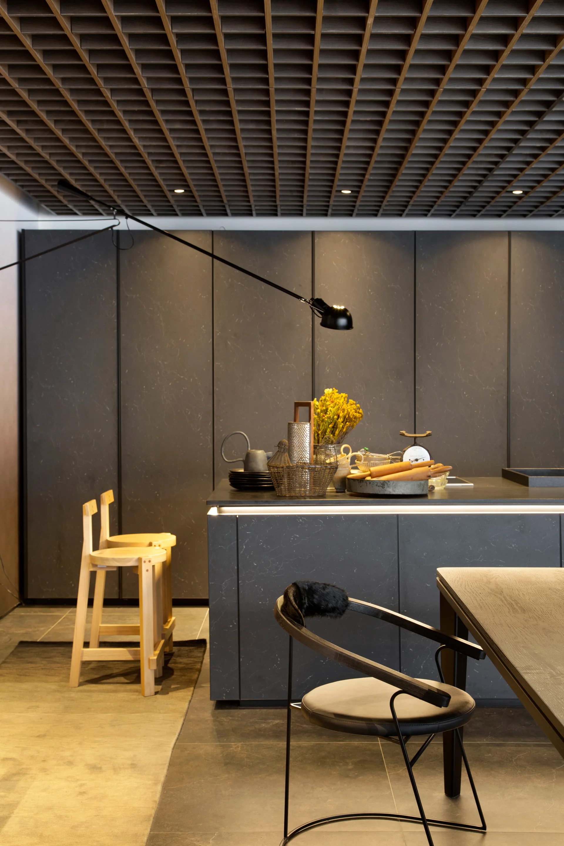 A modern kitchen featuring charcoal grey stone-like cabinetry, a central island with light wood bar stools, and a dark geometric grid ceiling.