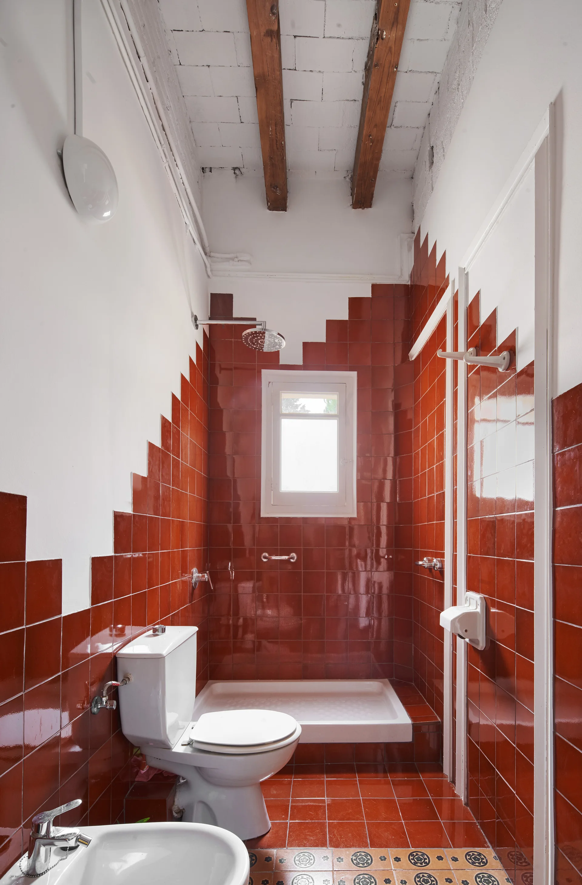 A narrow bathroom features glossy red wall tiles arranged in a stepped pattern, a white toilet, and exposed wooden ceiling beams.