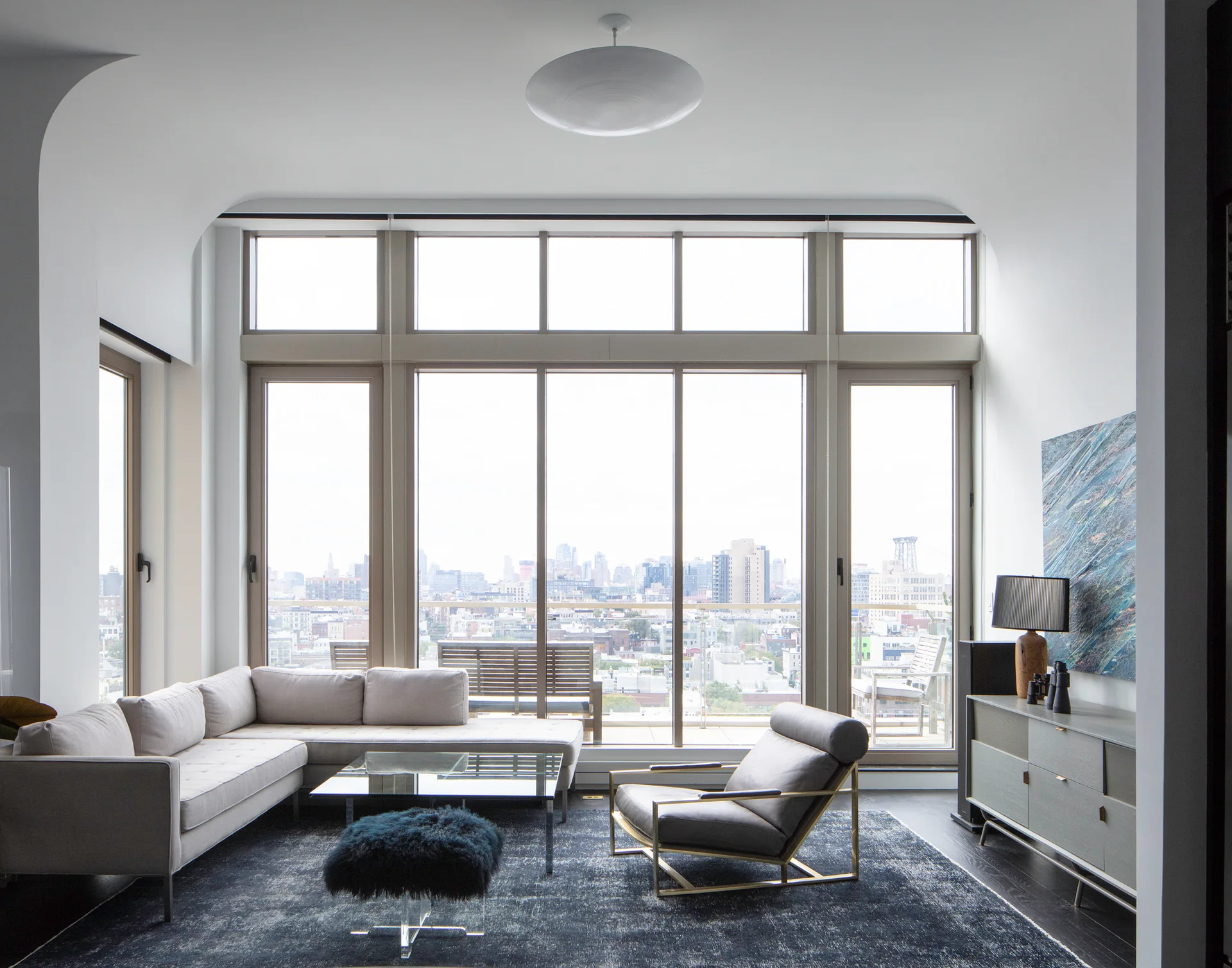 A modern living room featuring a grey sectional sofa, a leather armchair, a dark patterned rug, and large windows overlooking a city skyline.