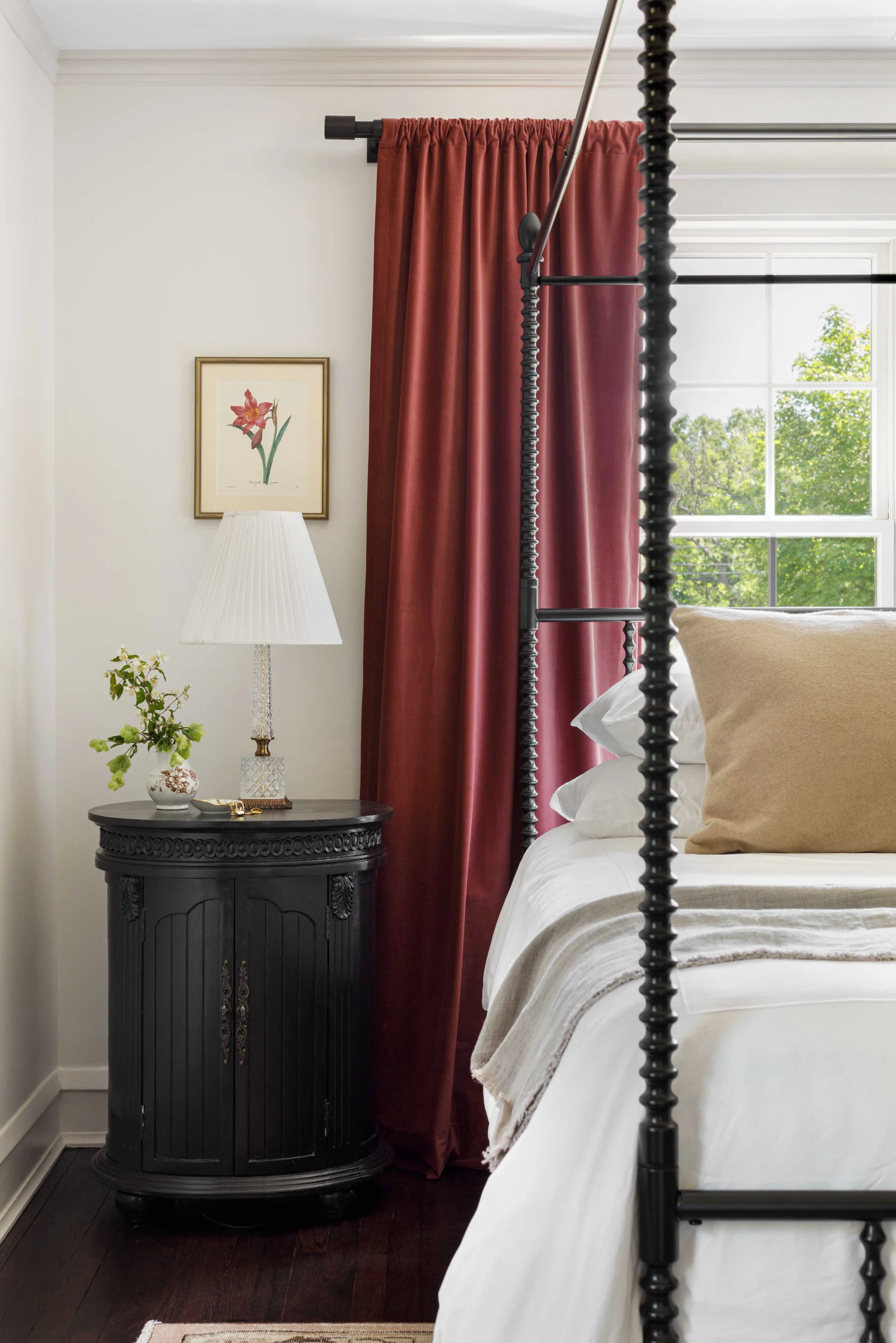 A black canopy bed with white linens sits next to a curved black wood nightstand topped with a pleated lamp in front of deep red curtains.