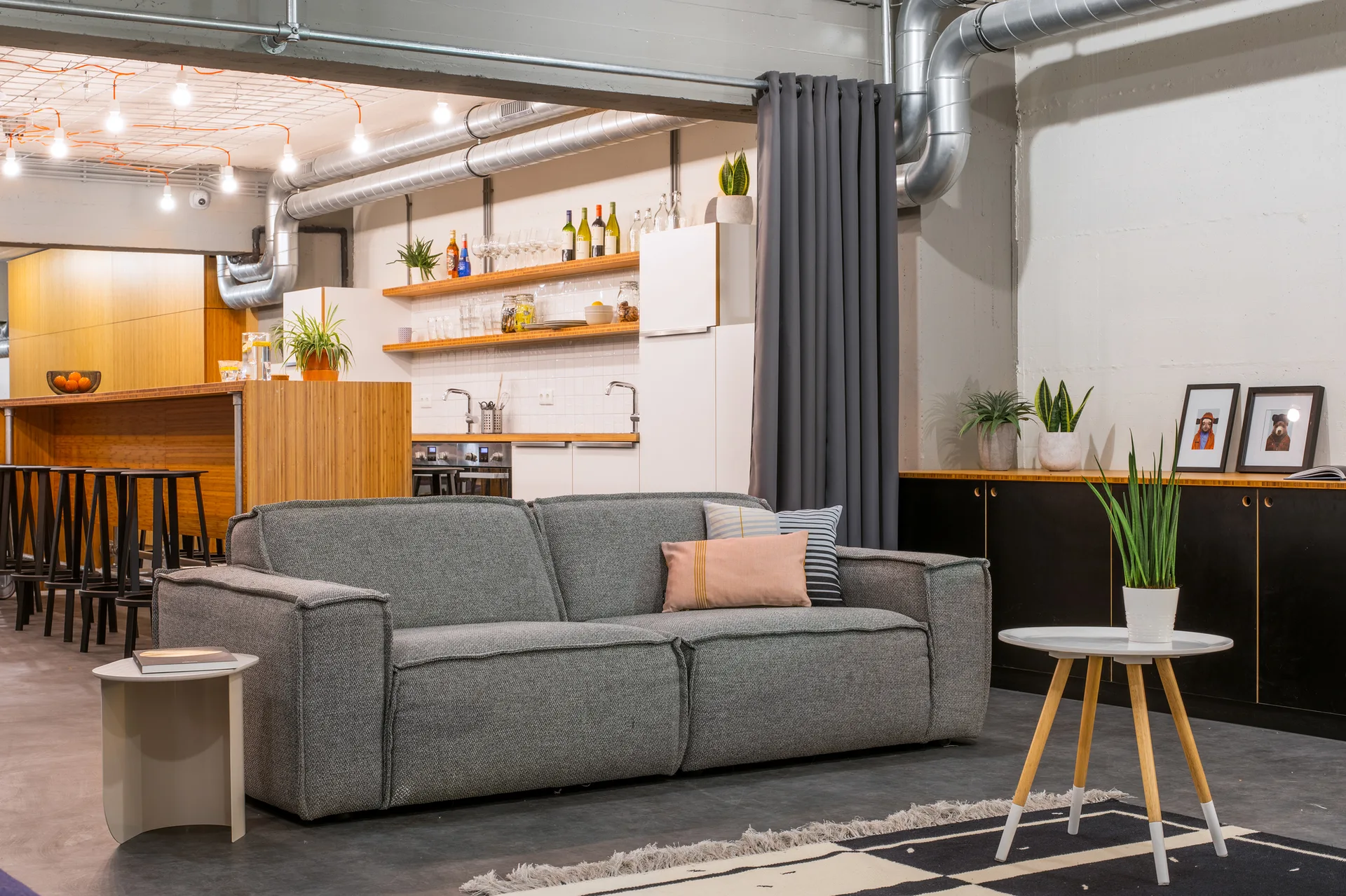 A grey sofa with throw pillows sits in an industrial living area featuring exposed ductwork, a dark storage cabinet, and an open kitchen.