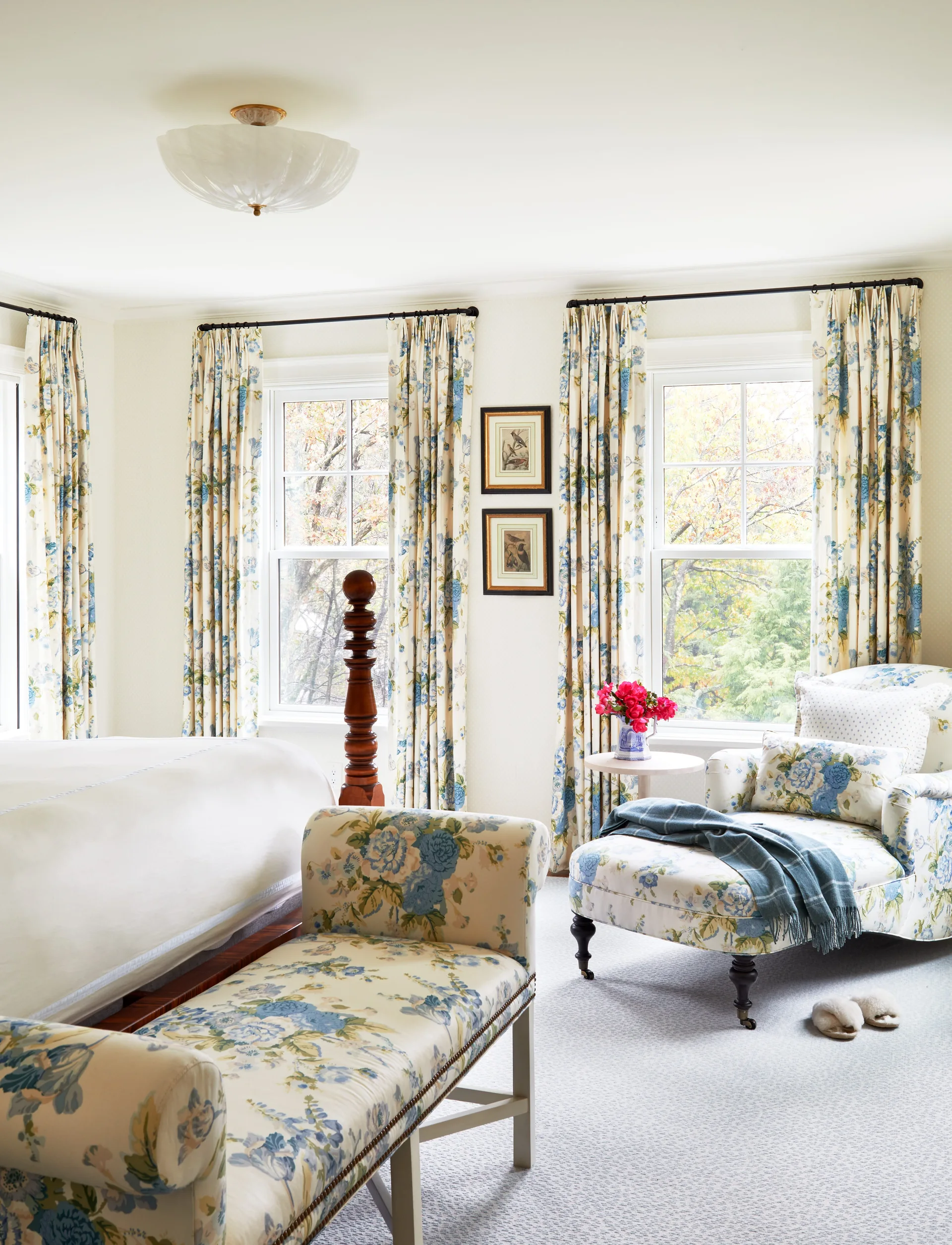 A traditional bedroom features a floral-upholstered chaise lounge and matching bench alongside windows with matching blue and yellow floral curtains.
