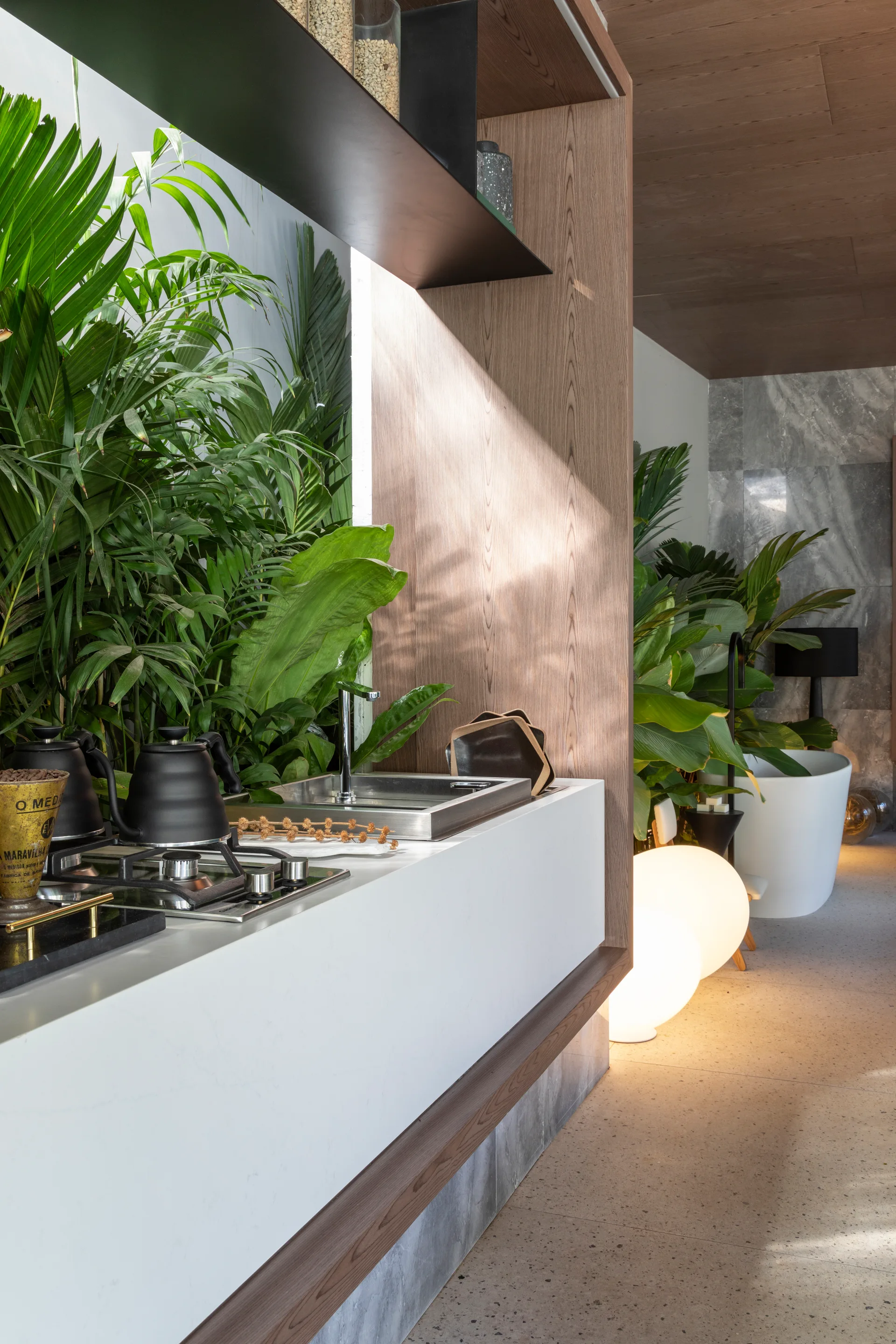 A modern white kitchen island with an integrated sink and stovetop is flanked by lush tropical houseplants and glowing spherical floor lamps.