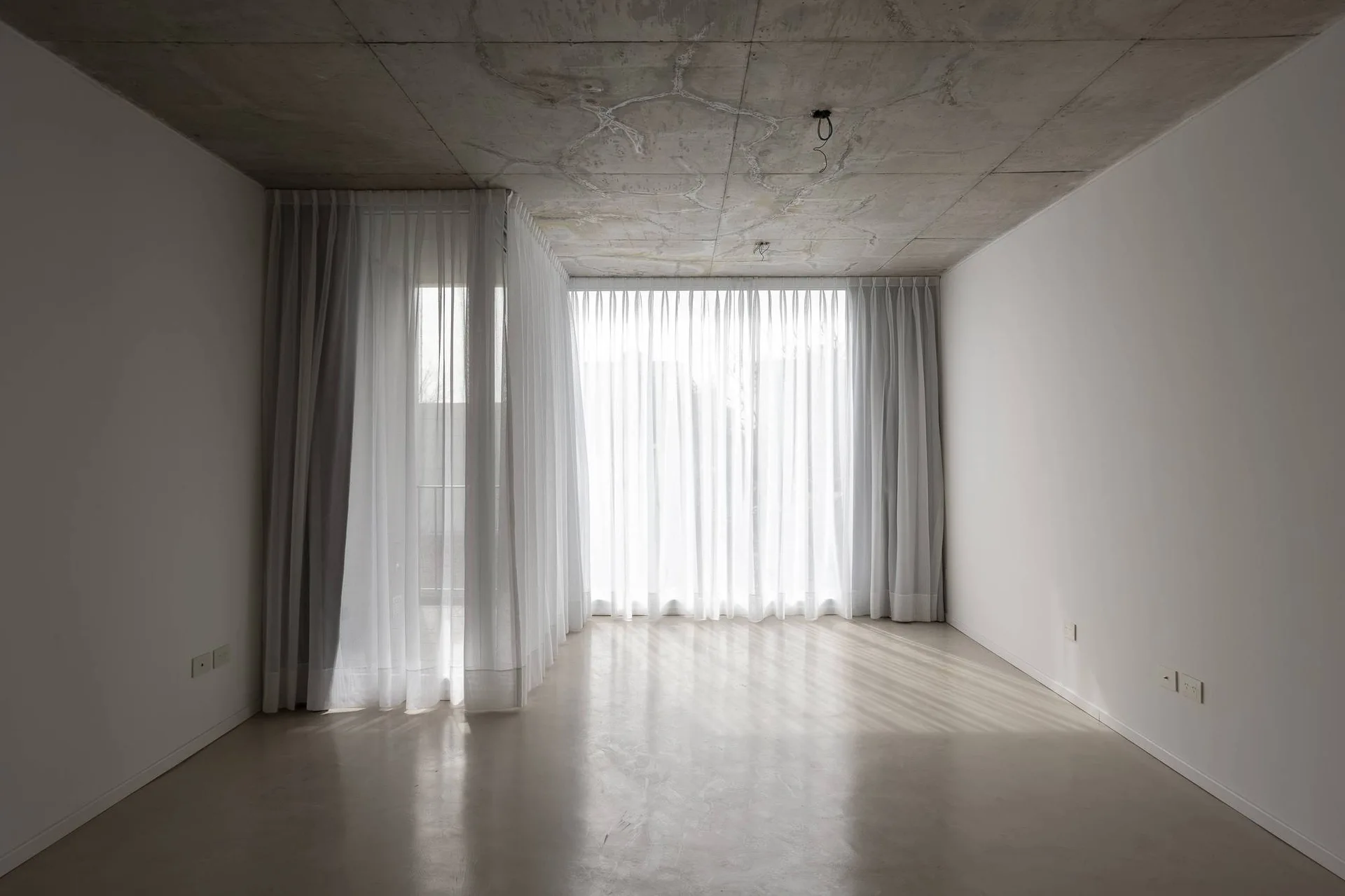 An empty minimalist room with a raw grey concrete ceiling, white walls, and floor-to-ceiling sheer white curtains filtering natural light.