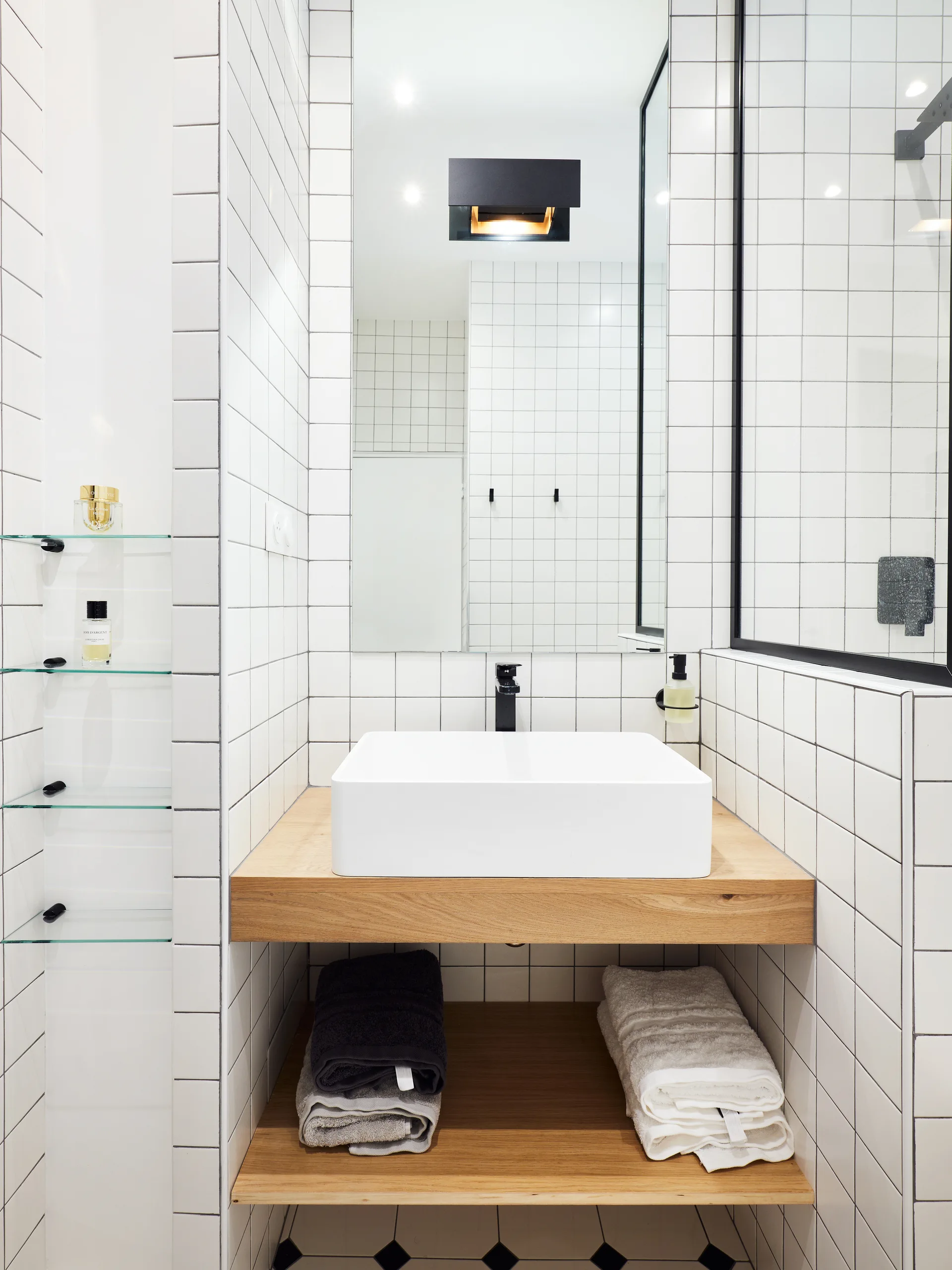 A modern bathroom features a white square vessel sink on a floating wooden vanity with an open shelf below, set against white grid wall tiles.