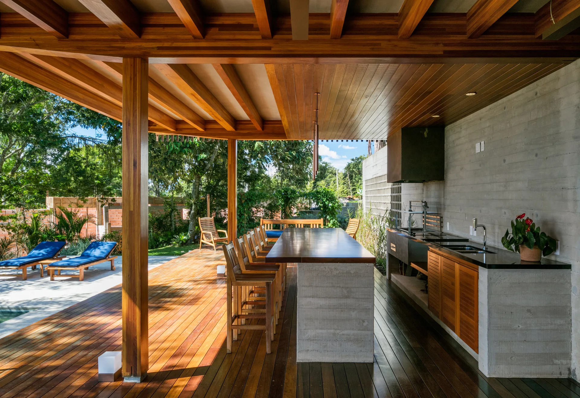 An outdoor kitchen with a concrete bar island, wooden stools, and timber ceiling beams sits on a wood patio next to a pool.
