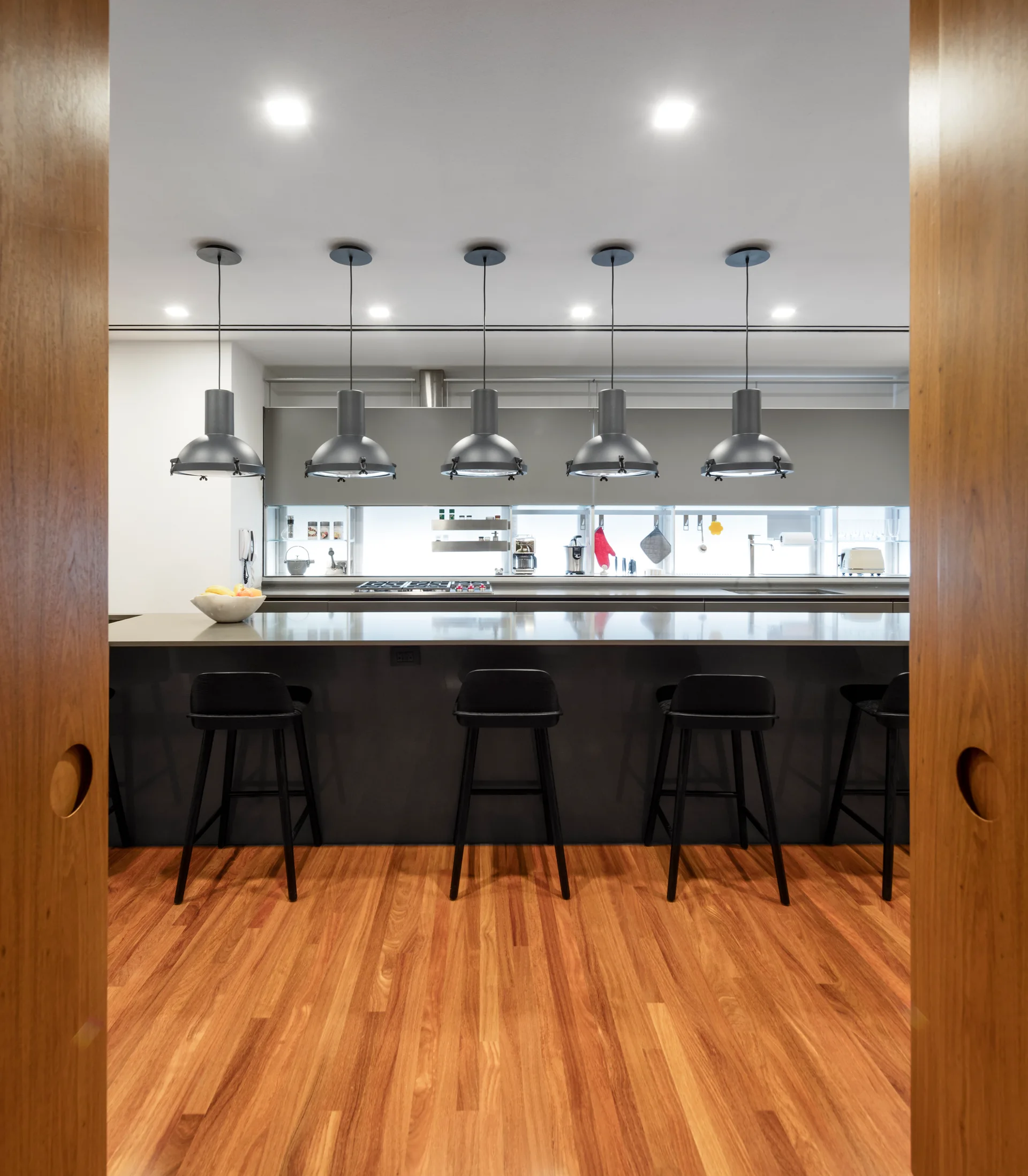 A modern kitchen viewed through wood doors, featuring a dark grey island, black bar stools, and five industrial pendant lights.