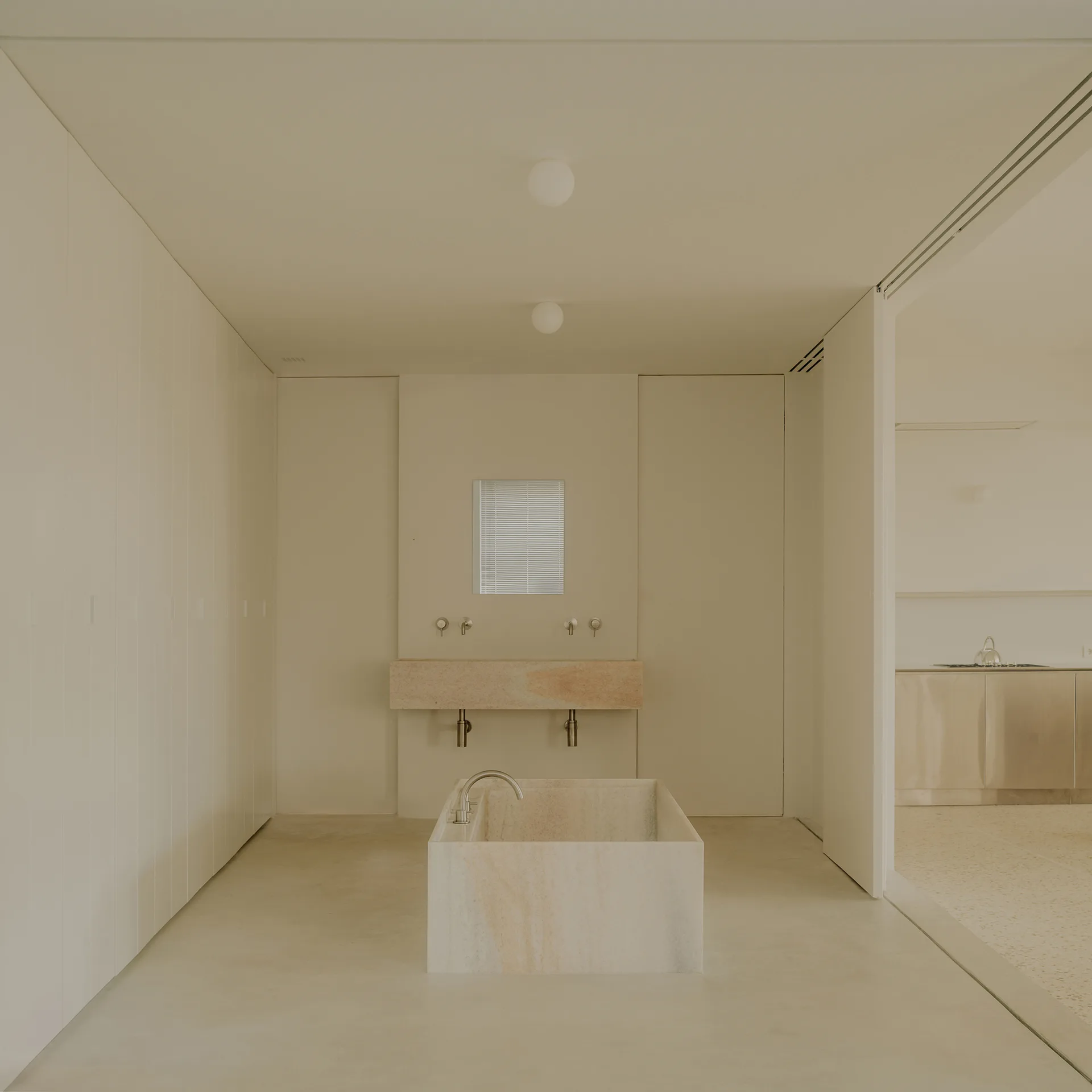 A minimalist cream bathroom featuring a central rectangular stone bathtub, a floating stone vanity, and flush full-height cabinetry.
