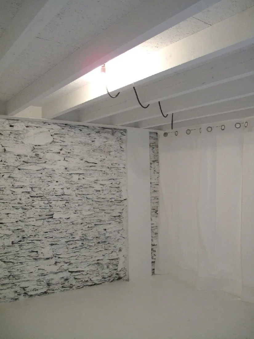 A minimalist room featuring a white painted textured stone wall, parallel ceiling beams, and a simple white fabric curtain divider.