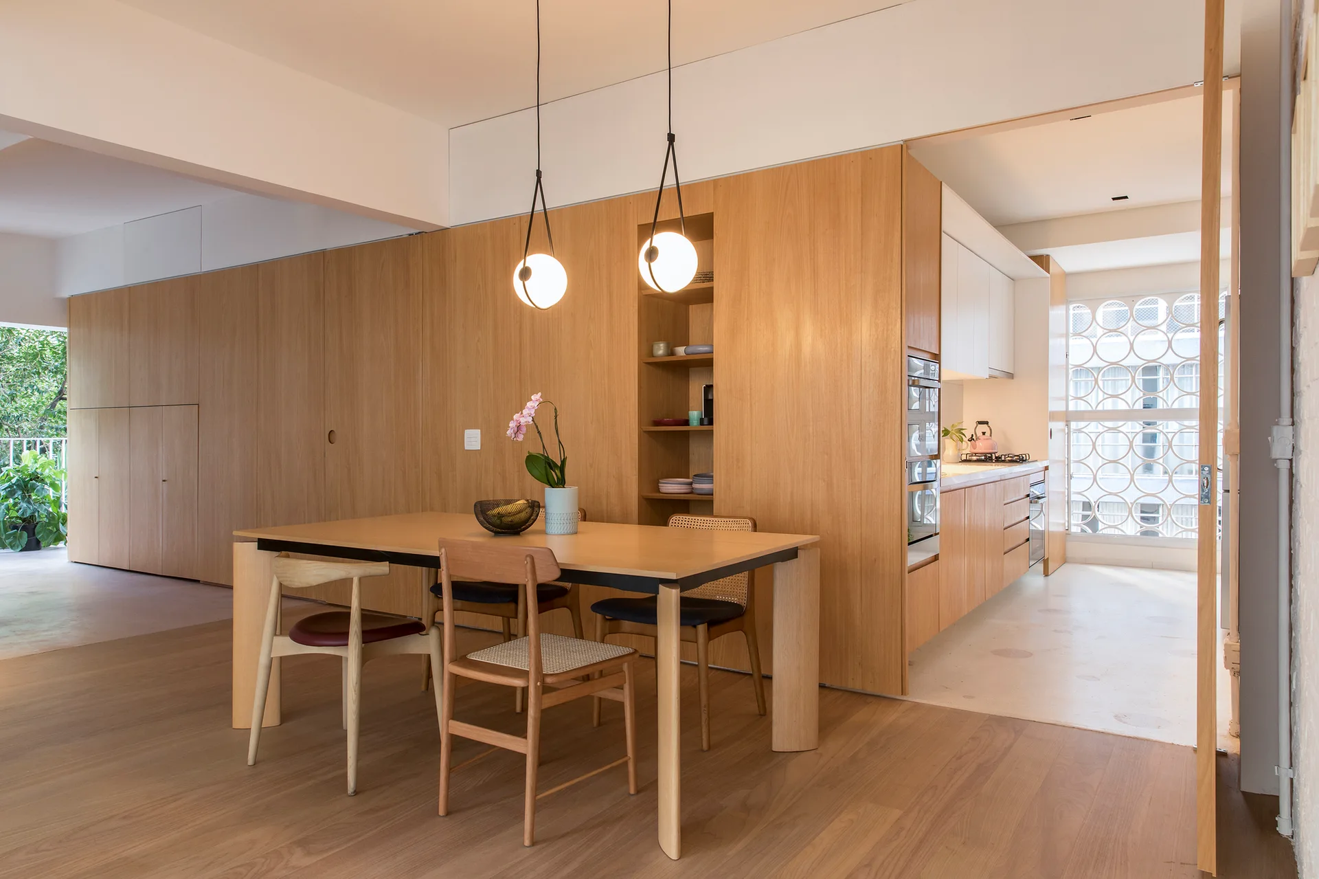 A modern dining room featuring a wooden table, mixed chairs, two hanging globe pendant lights, and a large wood-paneled storage wall.