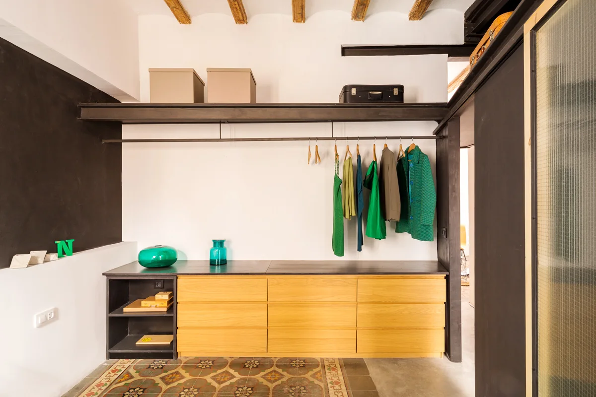 An open closet featuring a light wood drawer unit, a black metal hanging rack, and decorative tile flooring beneath exposed ceiling beams.