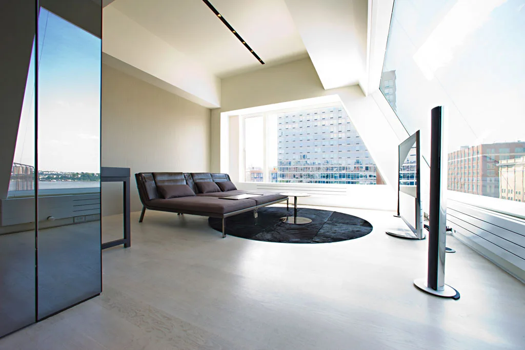 A minimalist living room features a low brown leather sofa on a round dark rug next to large angled floor-to-ceiling windows.
