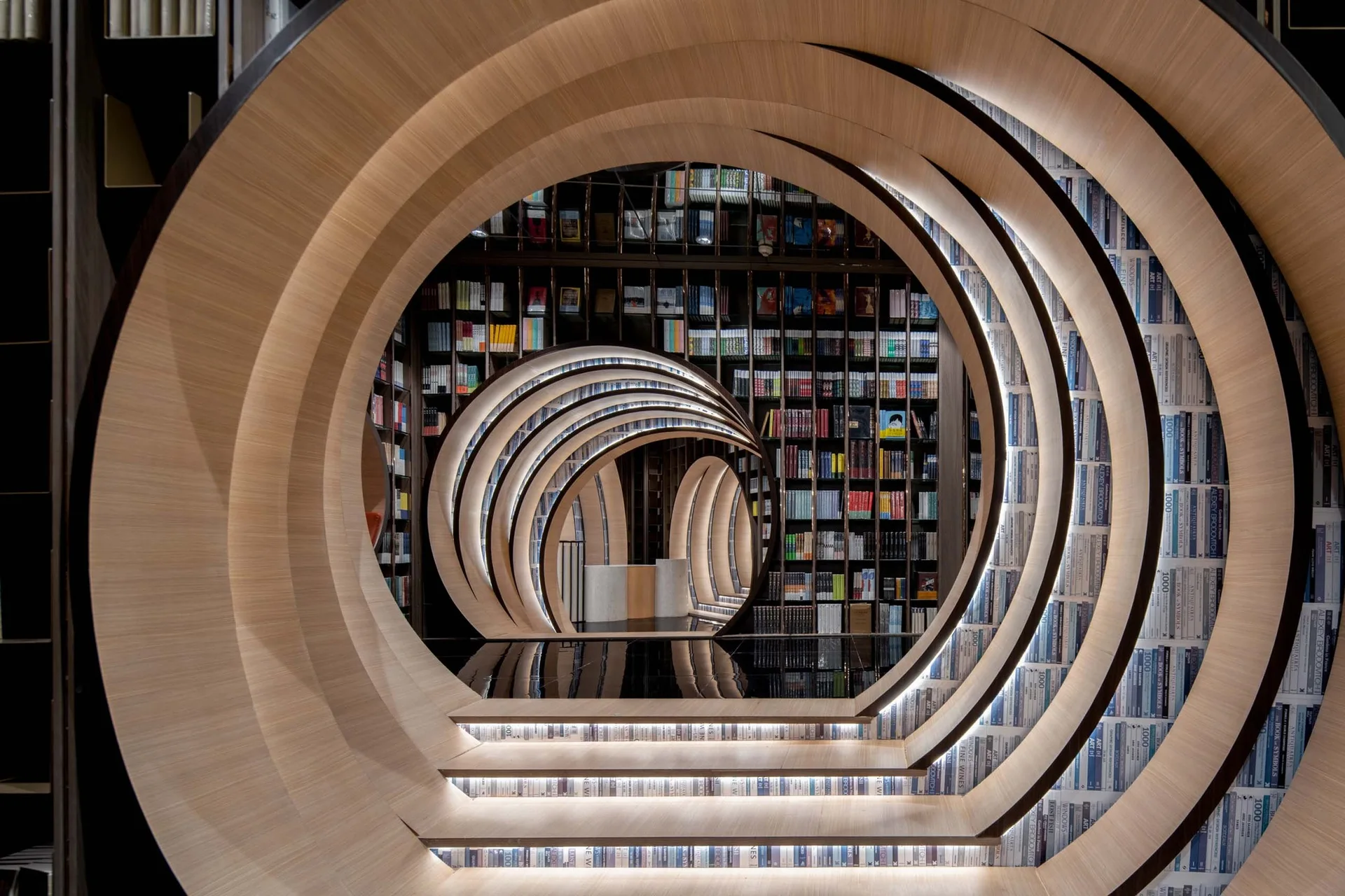 Concentric circular wooden archways frame a book-lined tunnel with illuminated steps and a glossy black floor.