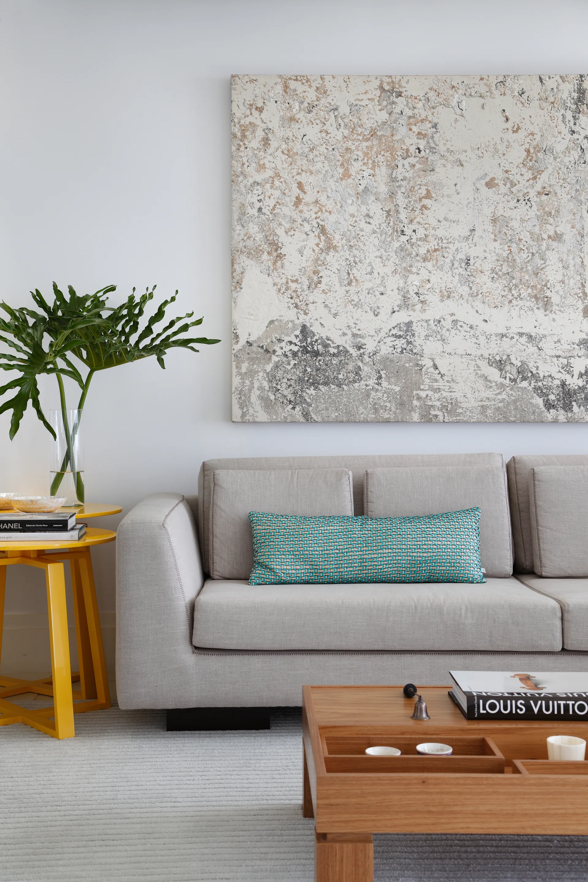 A beige fabric sofa with a patterned teal cushion sits beneath a large textured abstract painting next to a yellow side table and wooden coffee table.