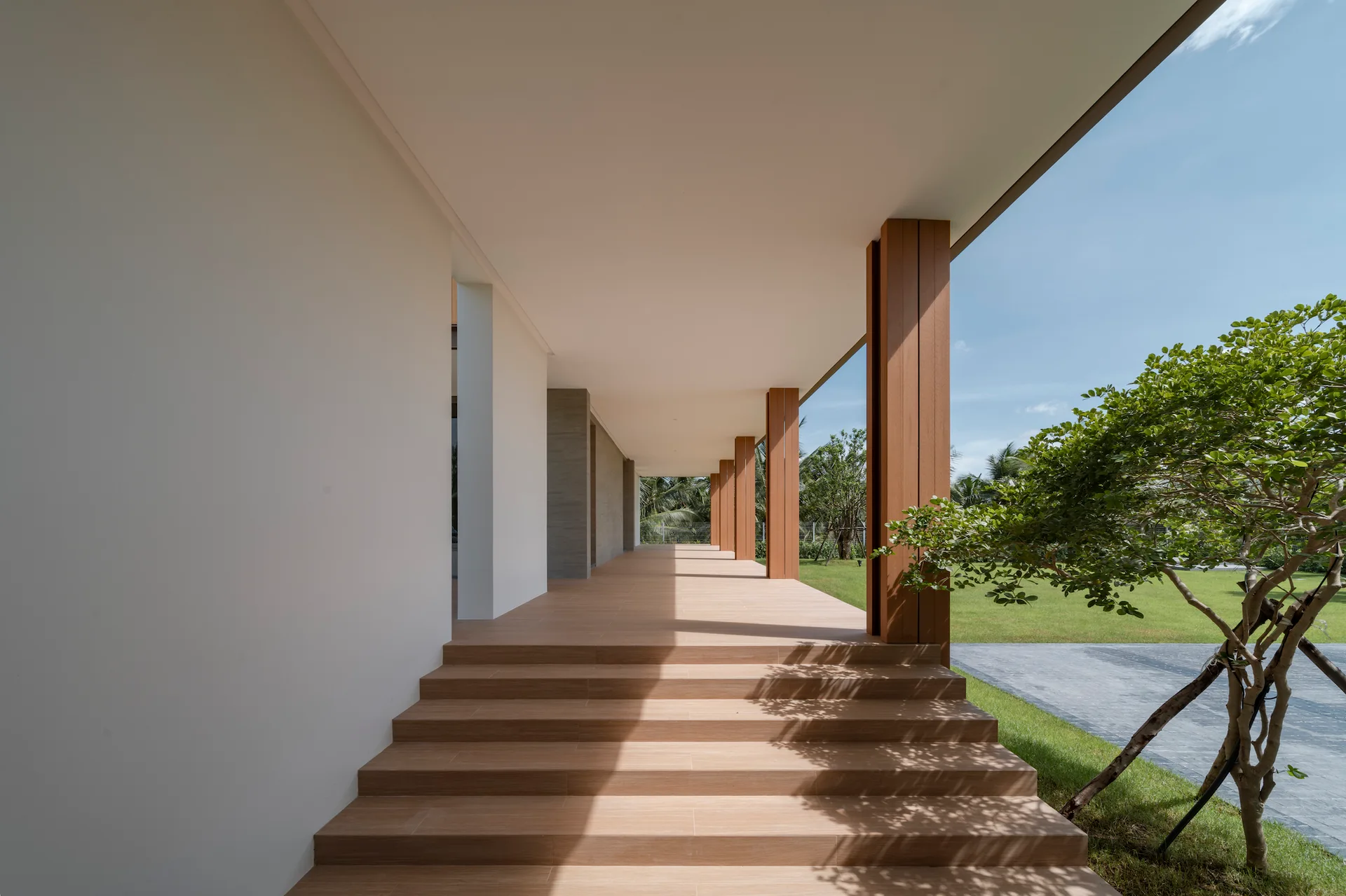 A minimalist covered terrace features wide wood-toned steps leading to a long walkway lined with square wooden pillars next to a green lawn.