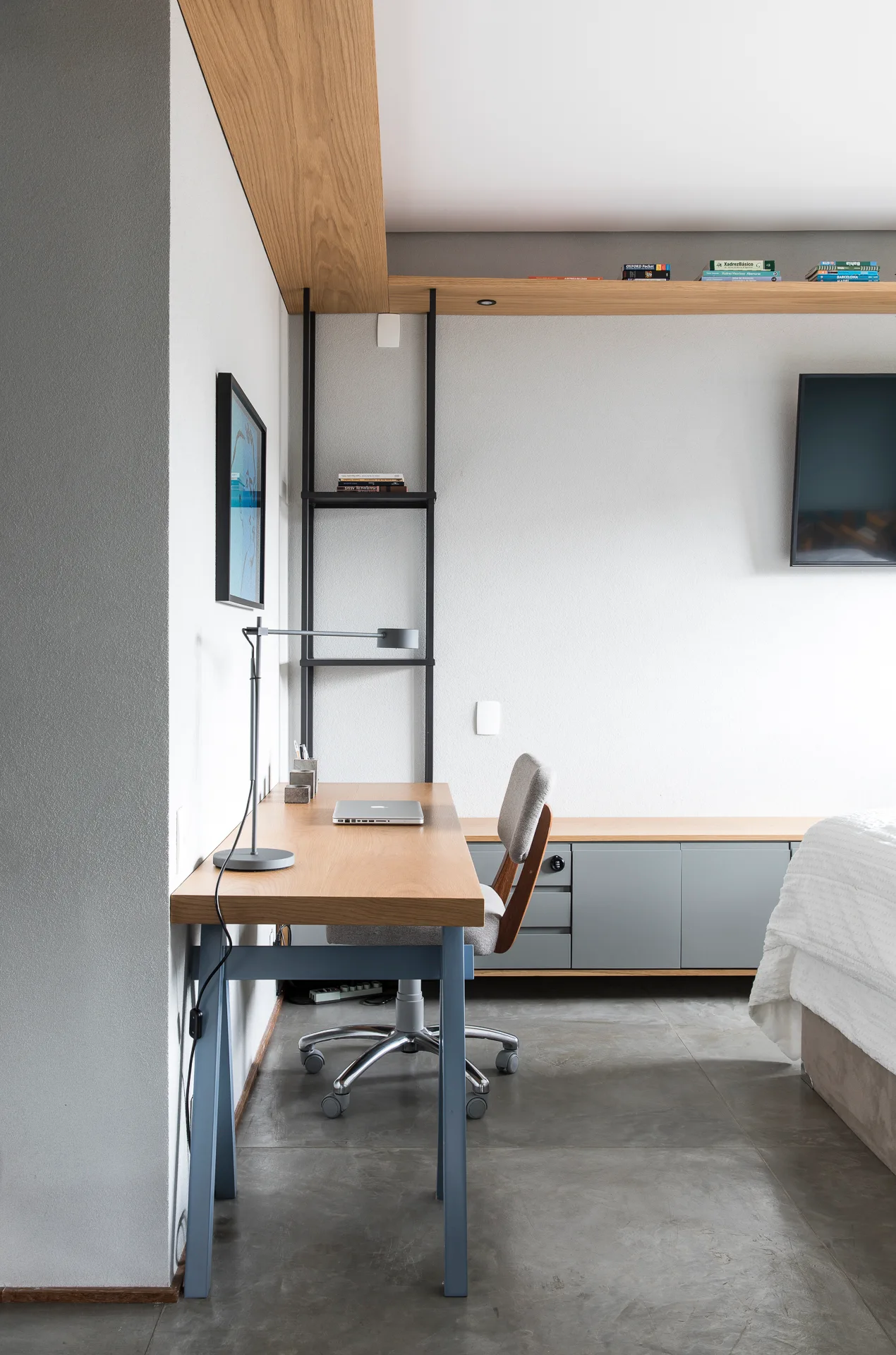 A modern bedroom workspace featuring a light wood desk, a grey office chair on wheels, and a long built-in wooden shelf above grey cabinets.