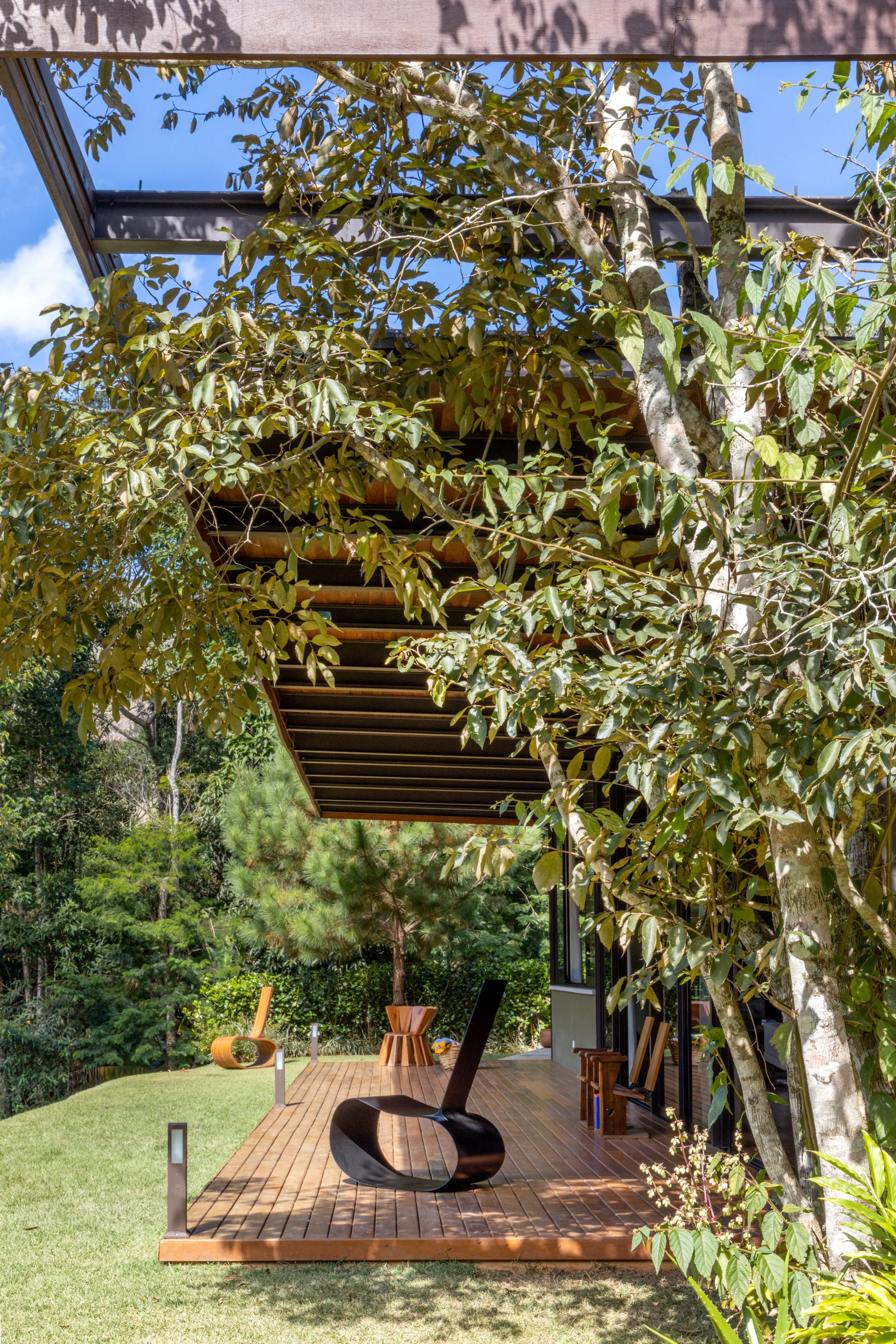 A modern black curved lounge chair sits on a slatted wooden deck under a tree-covered pergola next to a green lawn.