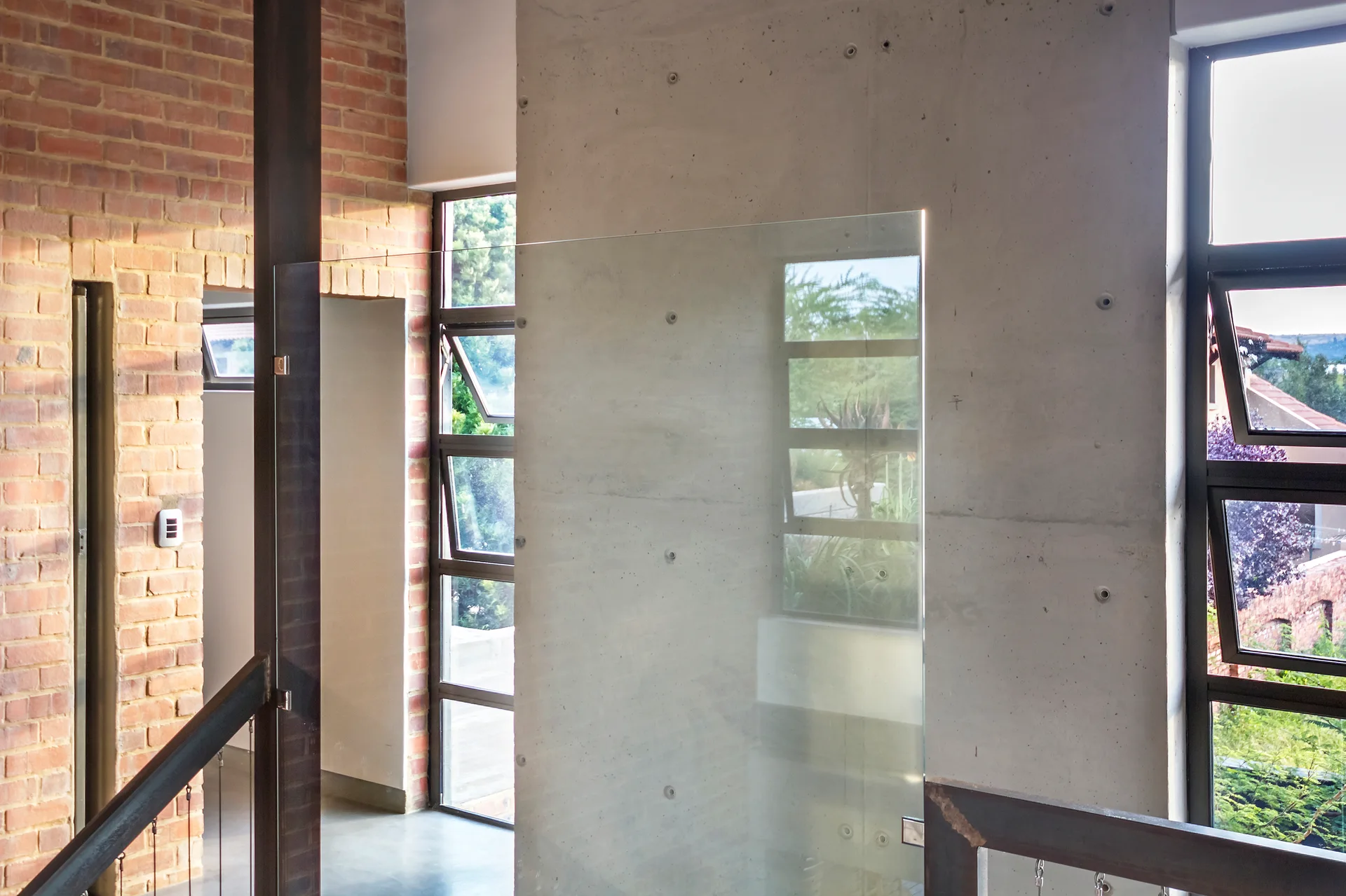 An industrial staircase landing features a raw concrete wall paneled with tie-rod holes next to an exposed red brick wall and dark-framed windows.