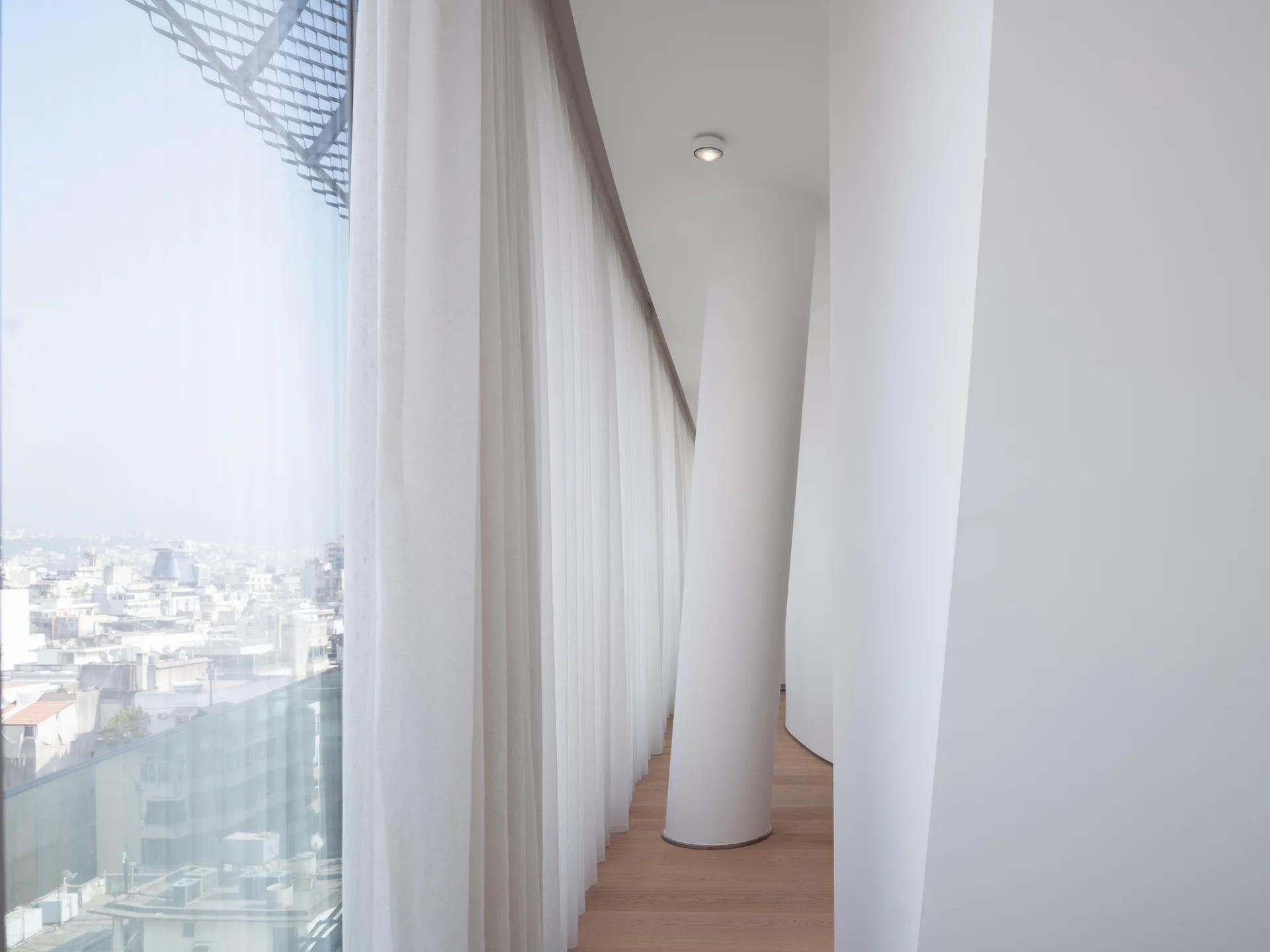 A minimalist white corridor features a slanted structural column, light wood floors, and floor-to-ceiling sheer curtains along a glass window wall.