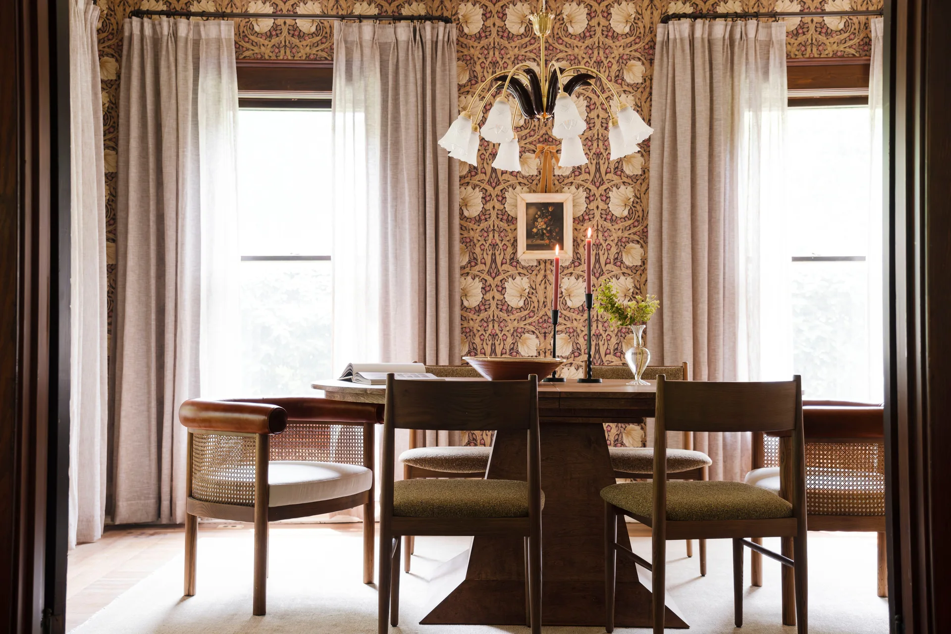 A wooden pedestal dining table and mismatched chairs sit in a dining room with floral wallpaper, sheer curtains, and a vintage chandelier.