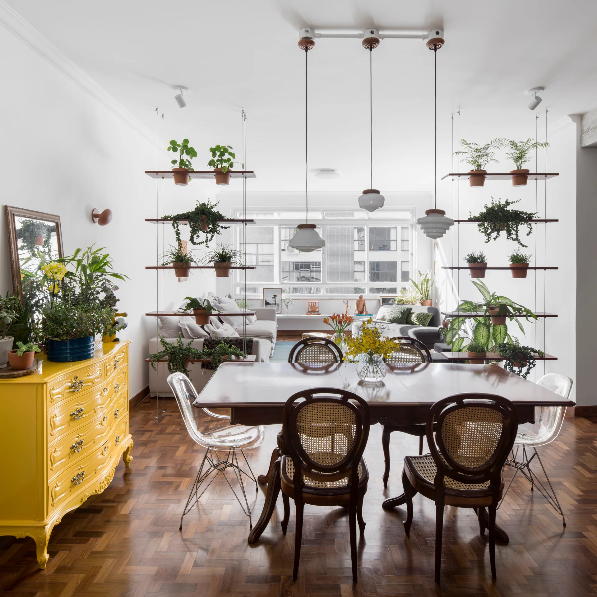 A dining room featuring a wooden table with cane and acrylic chairs, a bright yellow dresser, and hanging wooden shelves filled with potted plants.