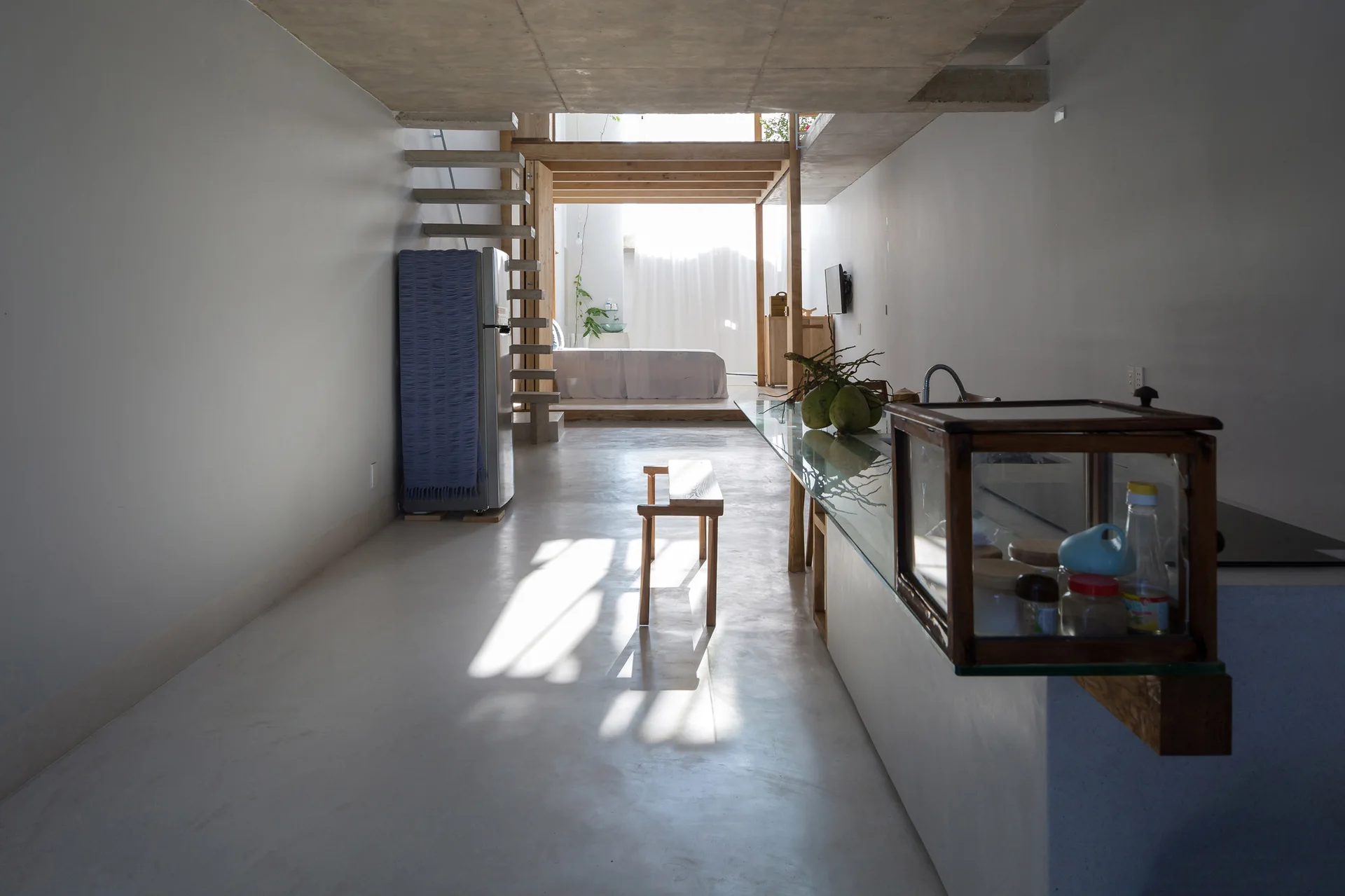 A minimalist narrow living space features concrete floors, an open concrete staircase, and a platform bed backlit by natural light.