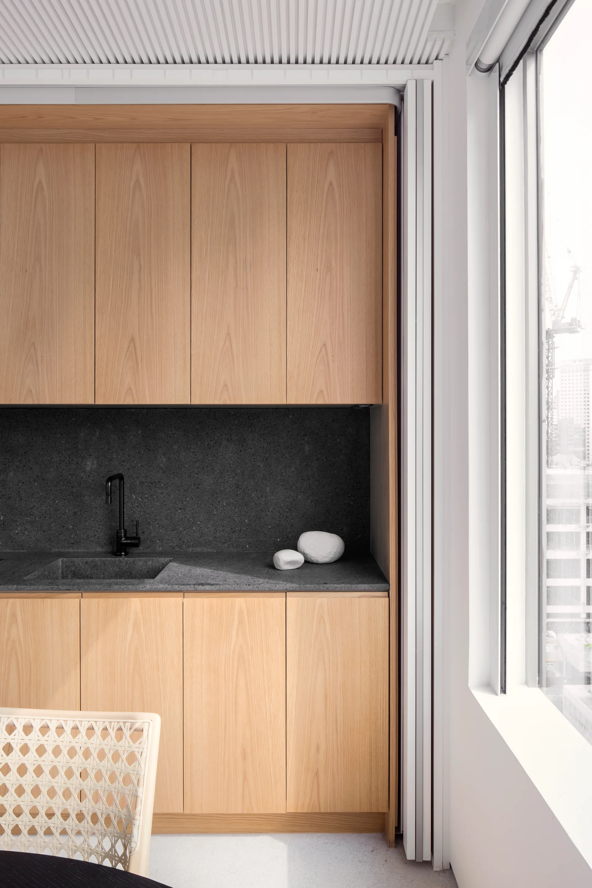 A minimalist kitchen corner features flat-front oak cabinets, a dark grey stone countertop with an integrated sink, and a black faucet next to a large window.
