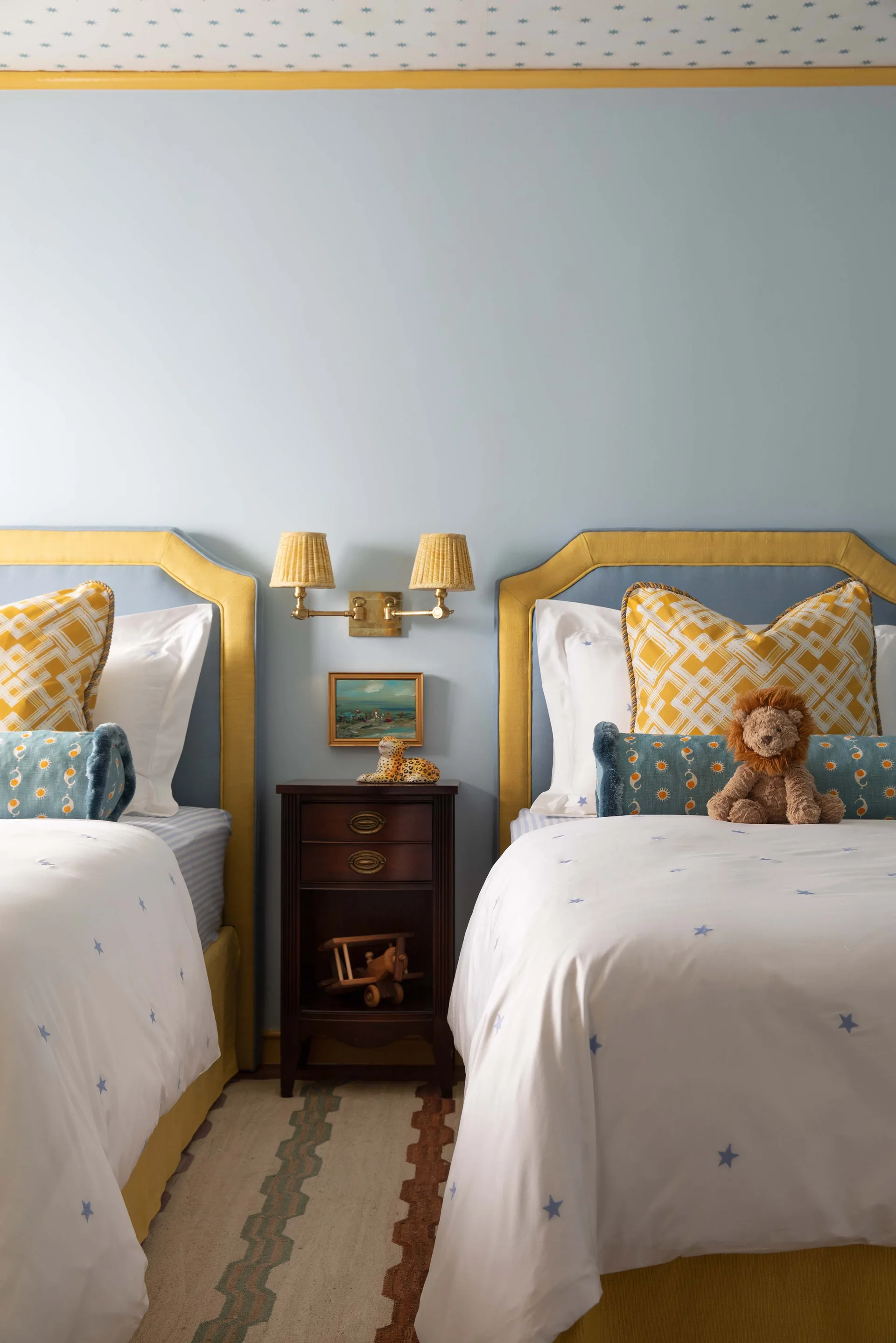 Two twin beds with yellow and blue upholstered headboards flank a dark wood nightstand in a child's bedroom with light blue walls.