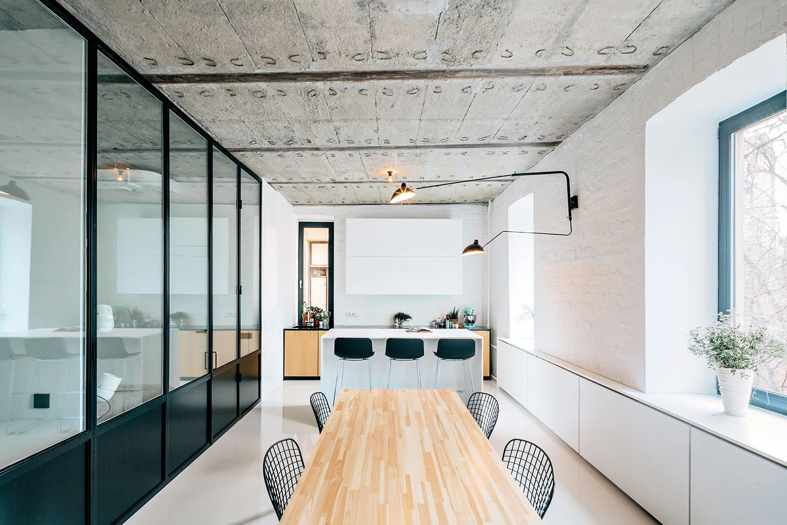 A modern dining room features a light wood table, wire chairs, a black metal glass partition, and an exposed concrete ceiling.