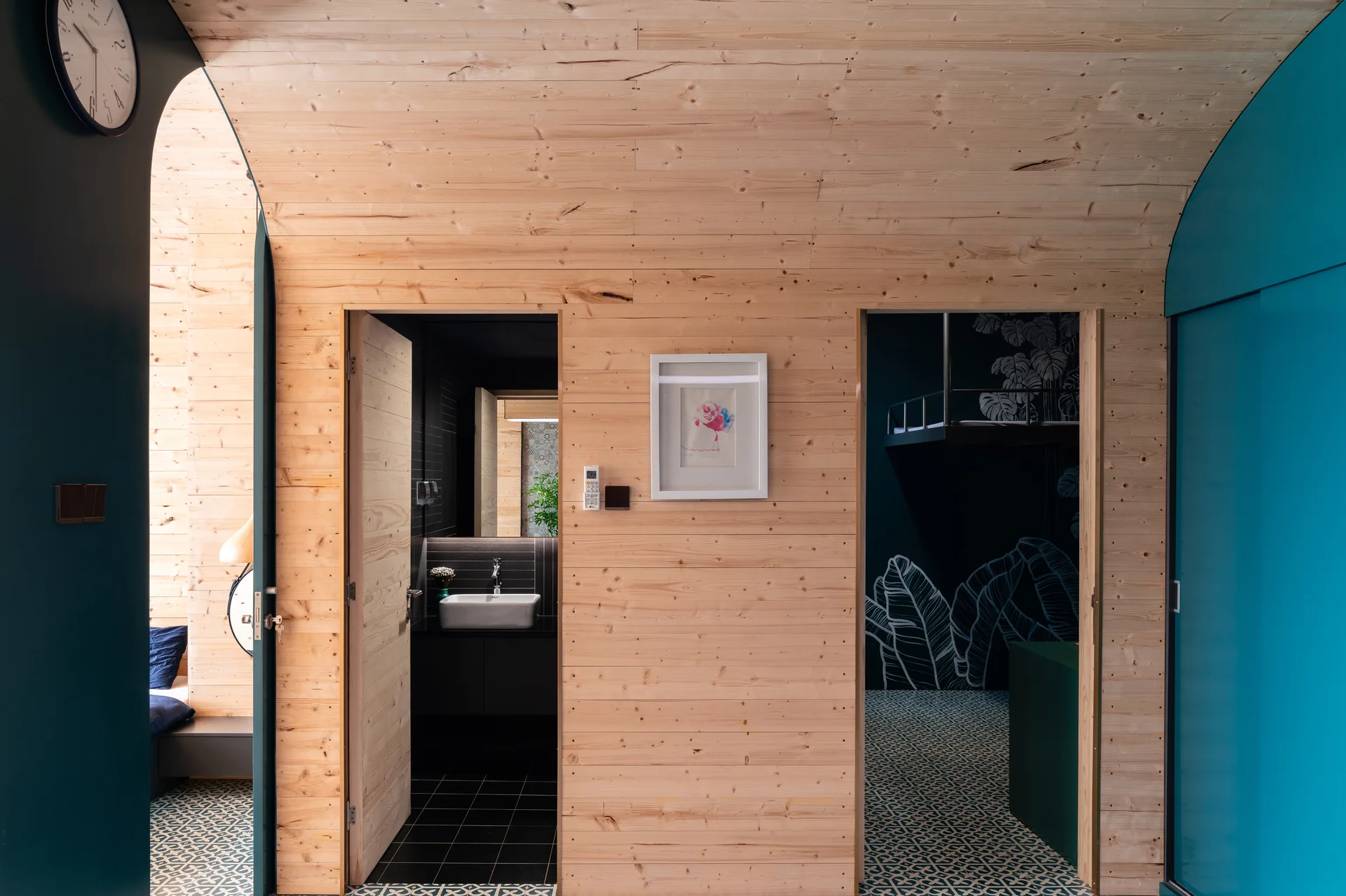 A wood-paneled hallway with a curved ceiling features two doorways showing a dark bathroom with a white sink and a bedroom with patterned tiles.