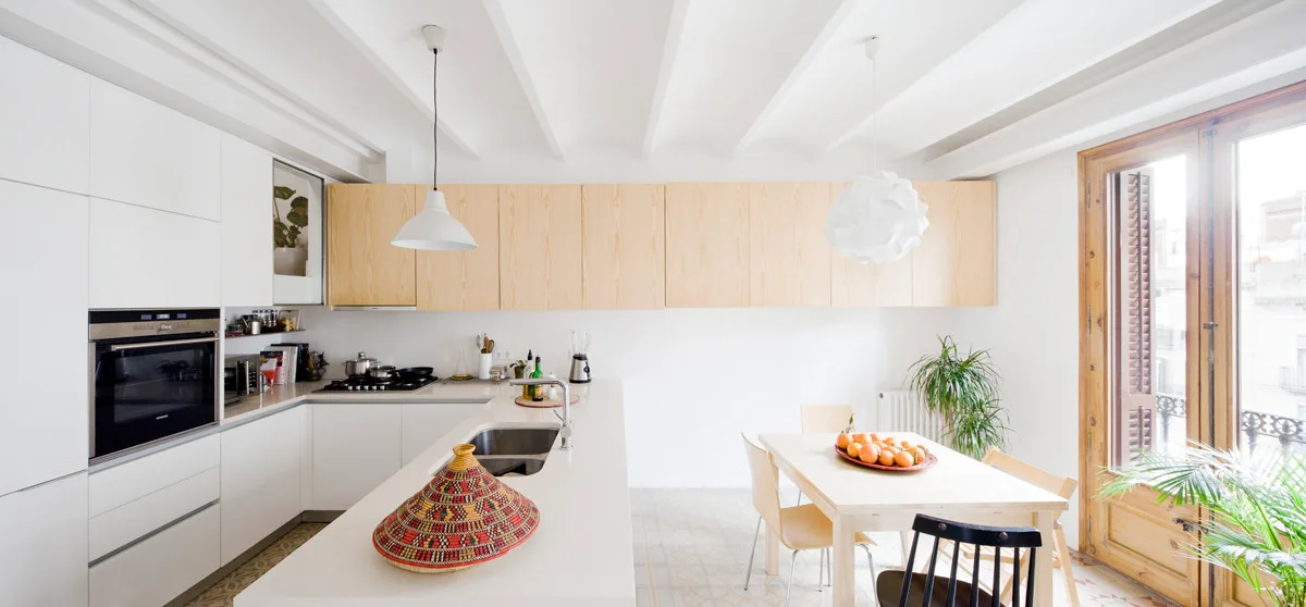 A bright minimalist kitchen featuring white cabinetry, a long block of light wood upper cupboards, exposed ceiling beams, and a wooden dining table.