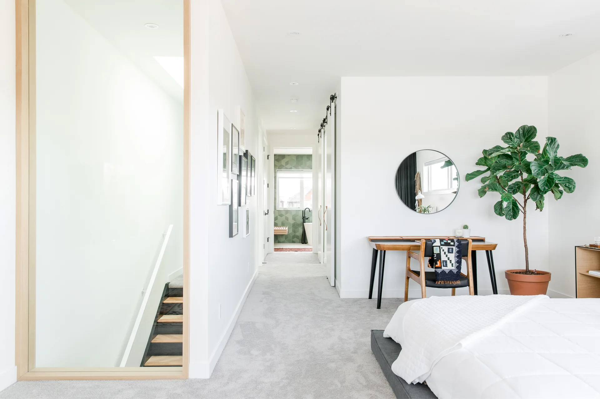 A bright white bedroom featuring a wood writing desk, a round mirror, a large fiddle leaf fig plant, and a view down a hallway to a tiled bathroom.