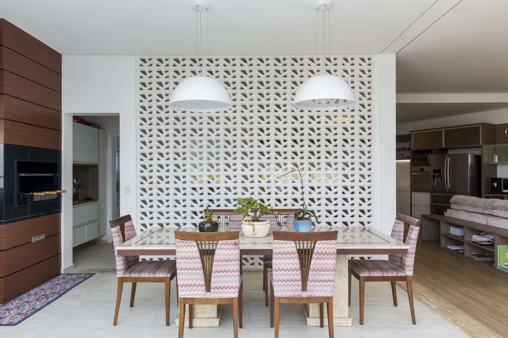 A dining area features a stone table and patterned chairs set against a white geometric breeze block partition wall under two dome pendant lights.