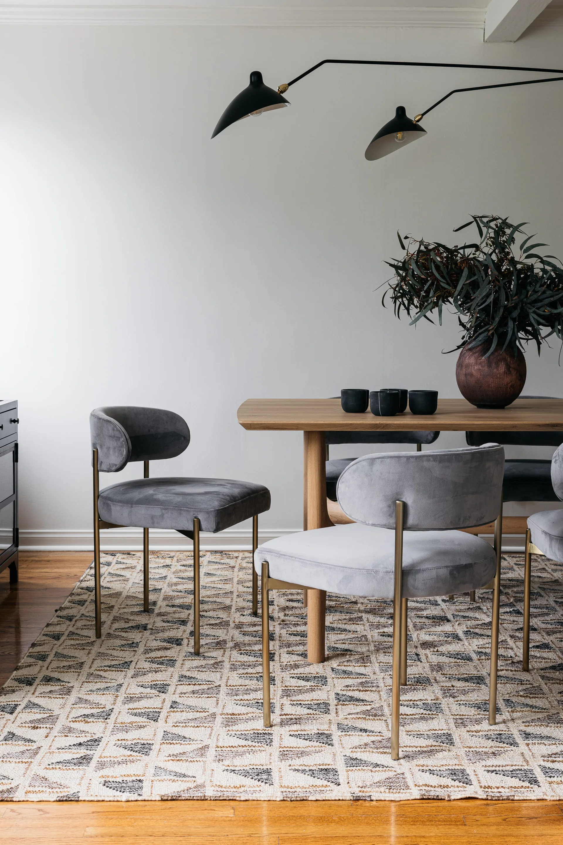 A wooden dining table is surrounded by grey upholstered chairs with brass legs on a geometric patterned rug beneath a black multi-arm light fixture.