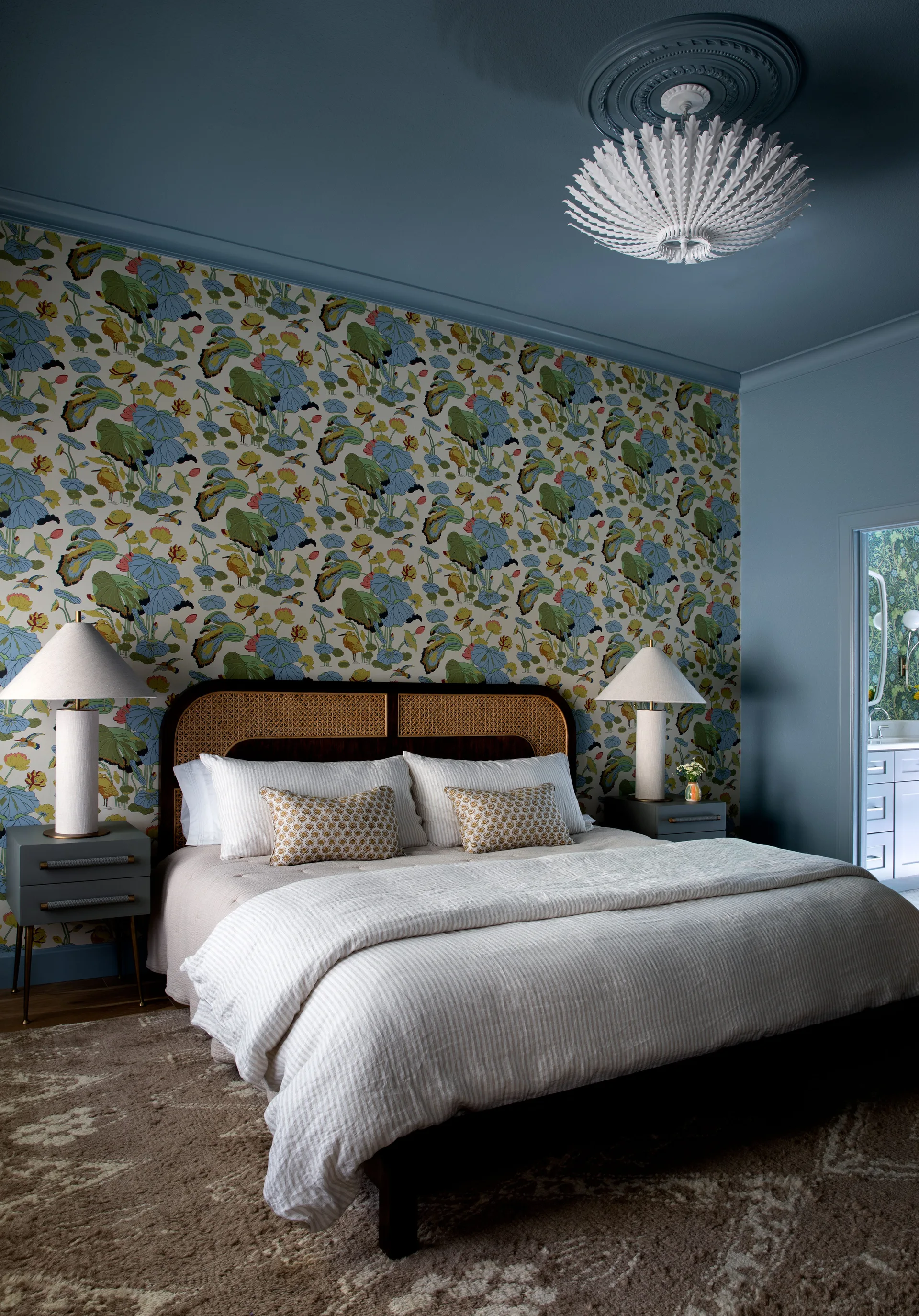 A bedroom featuring a bed with a woven cane headboard against botanical wallpaper, under a blue ceiling with a leaf-shaped light fixture.