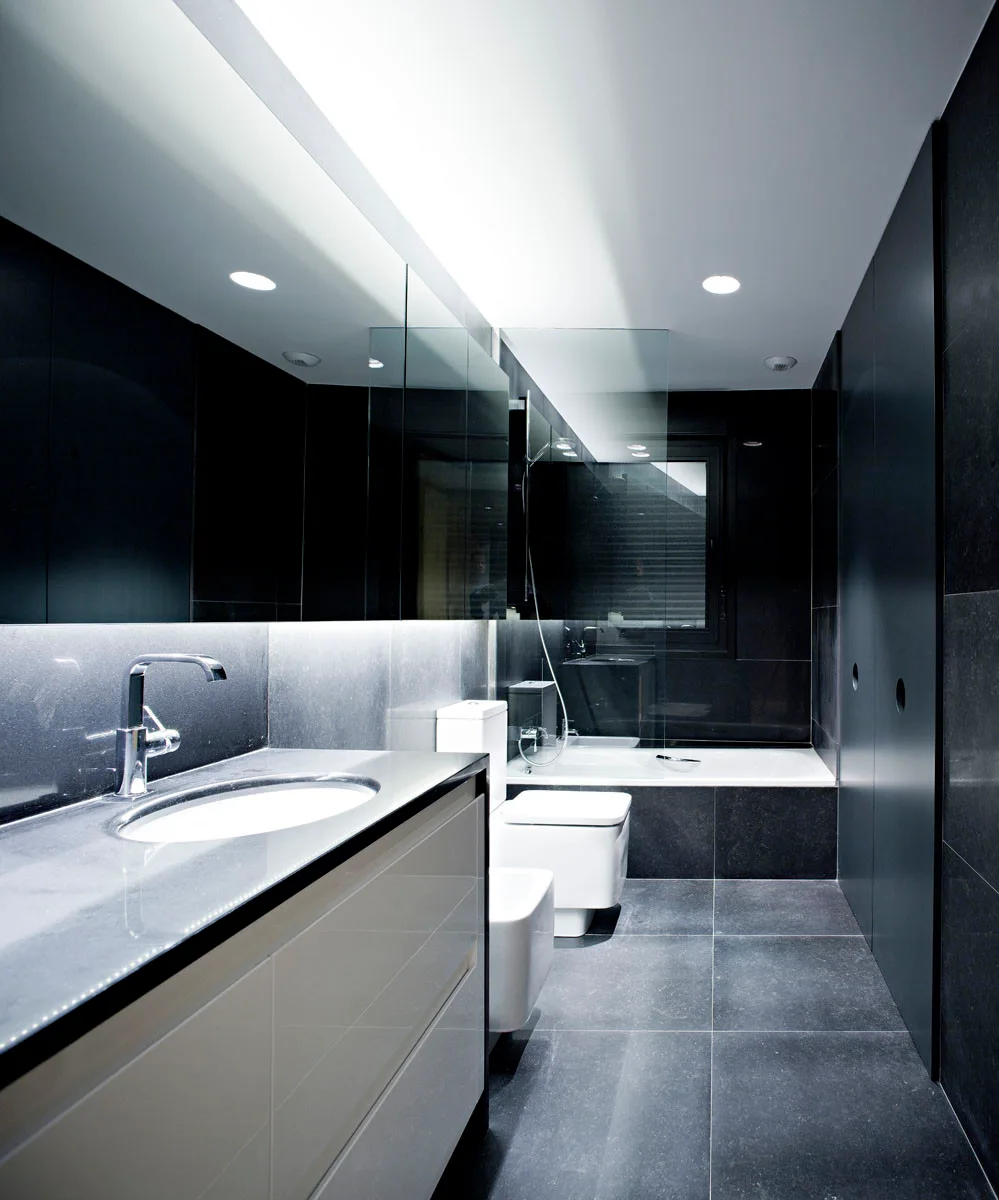 A minimalist modern bathroom features dark grey tile walls and floor, a white vanity with under-cabinet lighting, and a white toilet next to a bathtub.