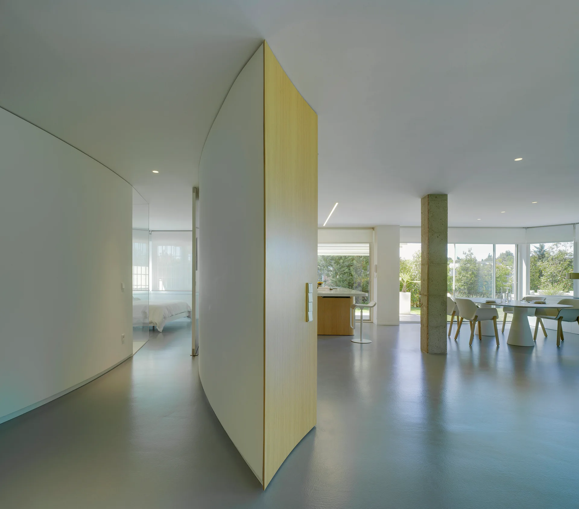 A modern open-plan interior with grey floors, a large curved wood partition wall, a dining area, and an exposed concrete column.